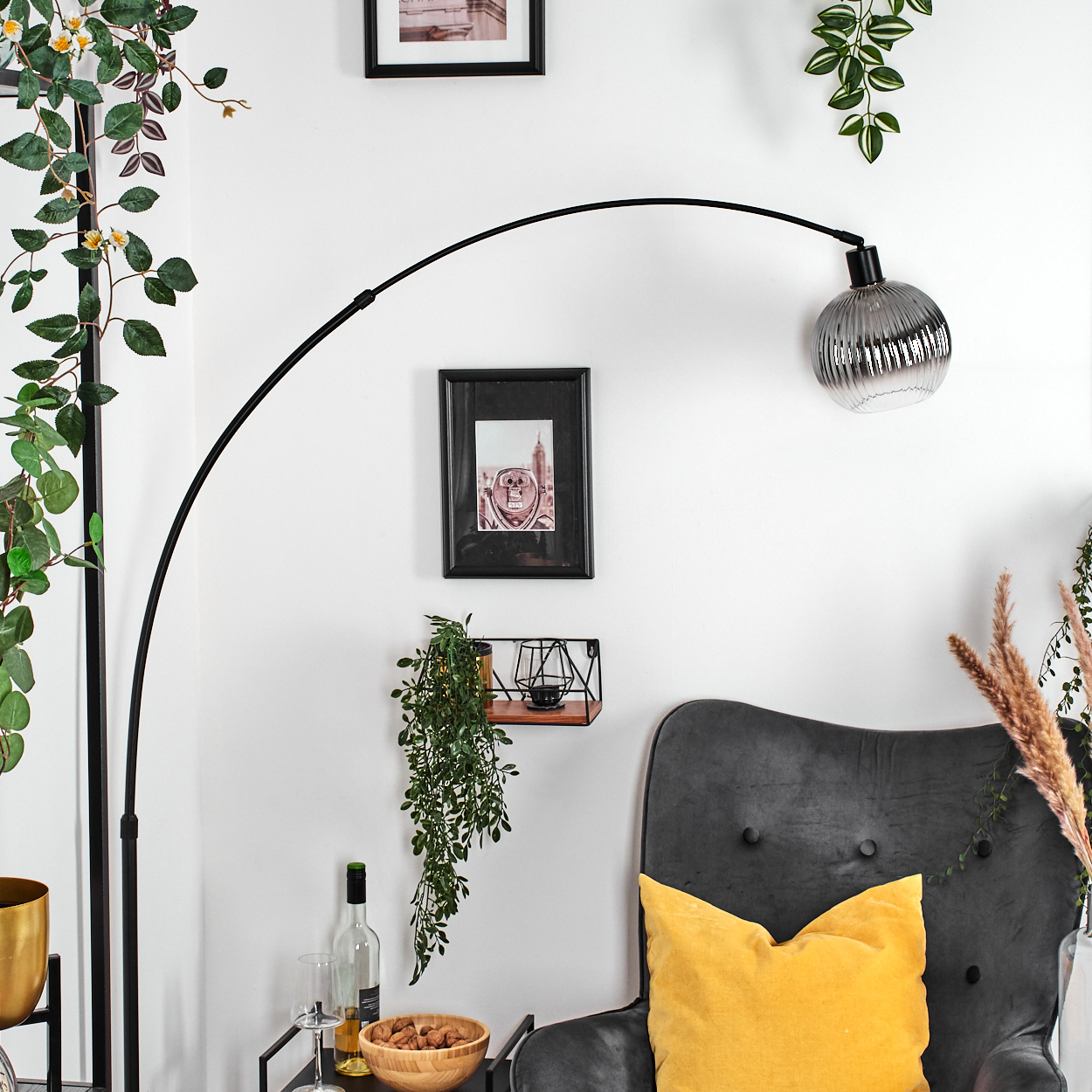 Ein Raum mit einer schwarzen Bogenlampe, gerahmten Bildern, einem grauen Sessel und Pflanzen.