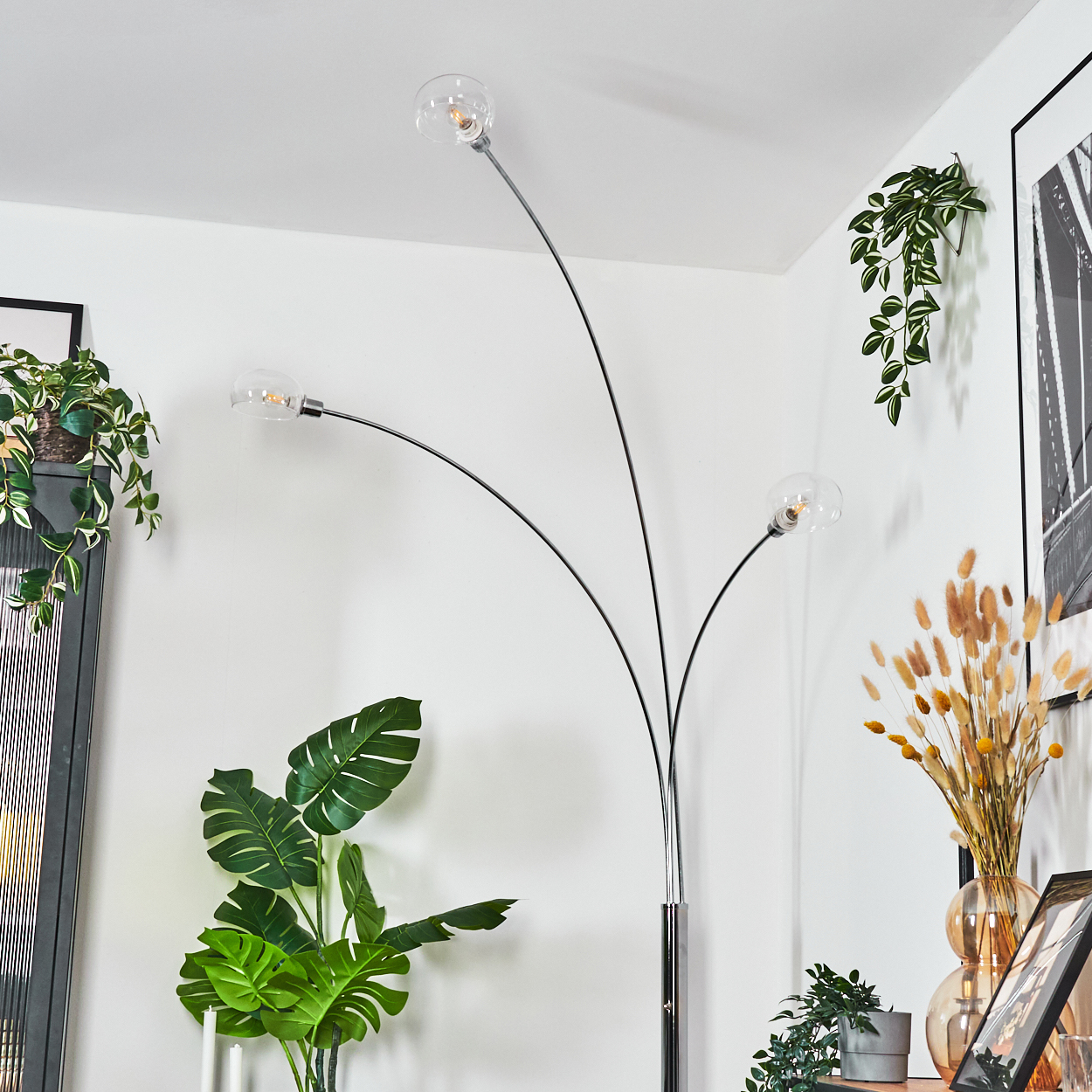 Ein modernes Zimmer mit einer hohen Bogenlampe mit drei kugelförmigen Glühbirnen, grünen Pflanzen und gerahmten Kunstwerken. Die Szene ist hell und luftig.