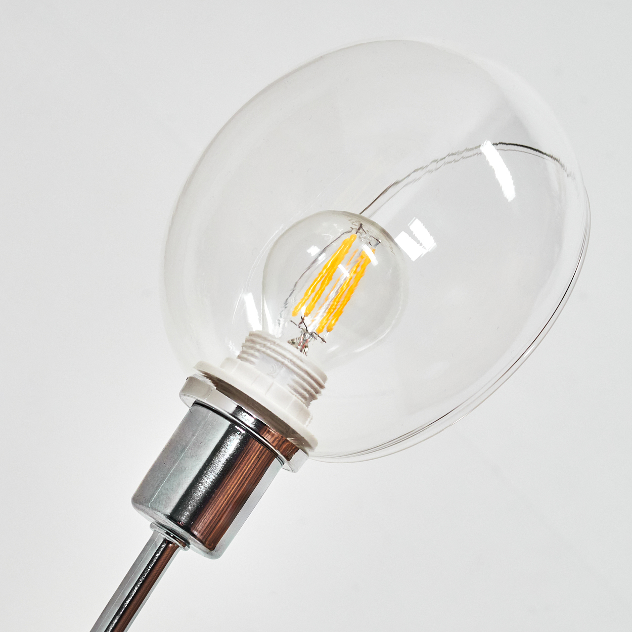 Nahaufnahme einer Lampe. Eine klare Glaskugel umschließt eine kleinere Glühbirne mit gelben Filamenten. Chromarmatur und weißer Hintergrund.