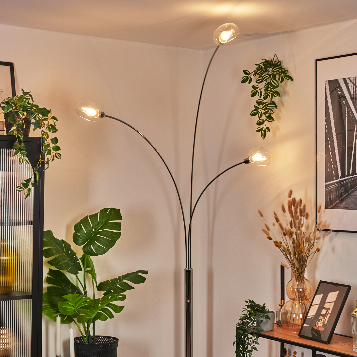 Eine moderne Stehlampe mit vier Glühbirnen steht in einer Ecke. Pflanzen und Bilder schmücken den Raum. Warmes Licht erhellt die Szene.