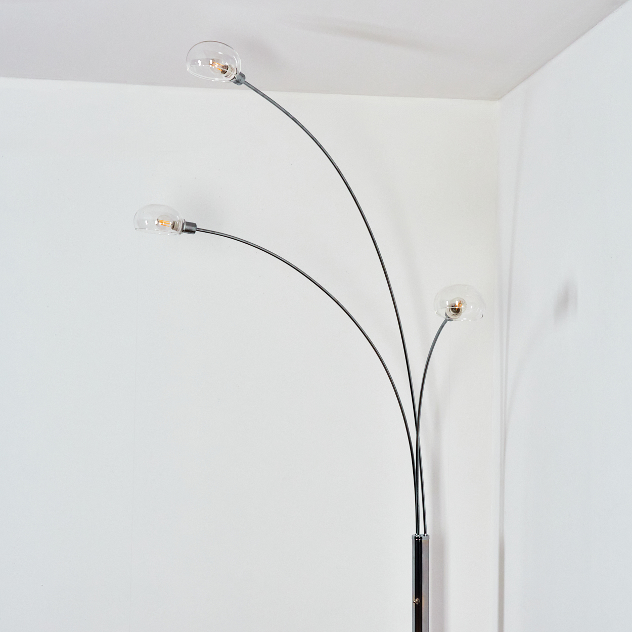 Eine moderne Stehlampe mit drei gebogenen Armen, die jeweils eine klare, kugelförmige Glühbirne halten. Die Lampe steht vor einer weißen Wand.