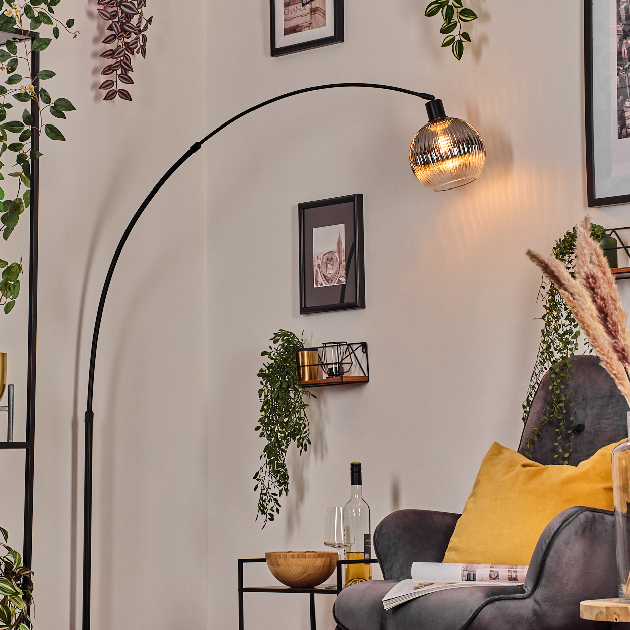 Ein modernes Wohnzimmer mit schwarzer Bogenlampe, grauem Sessel, gelbem Kissen, Pflanzen, Bildern und einem Beistelltisch mit Dekoration.