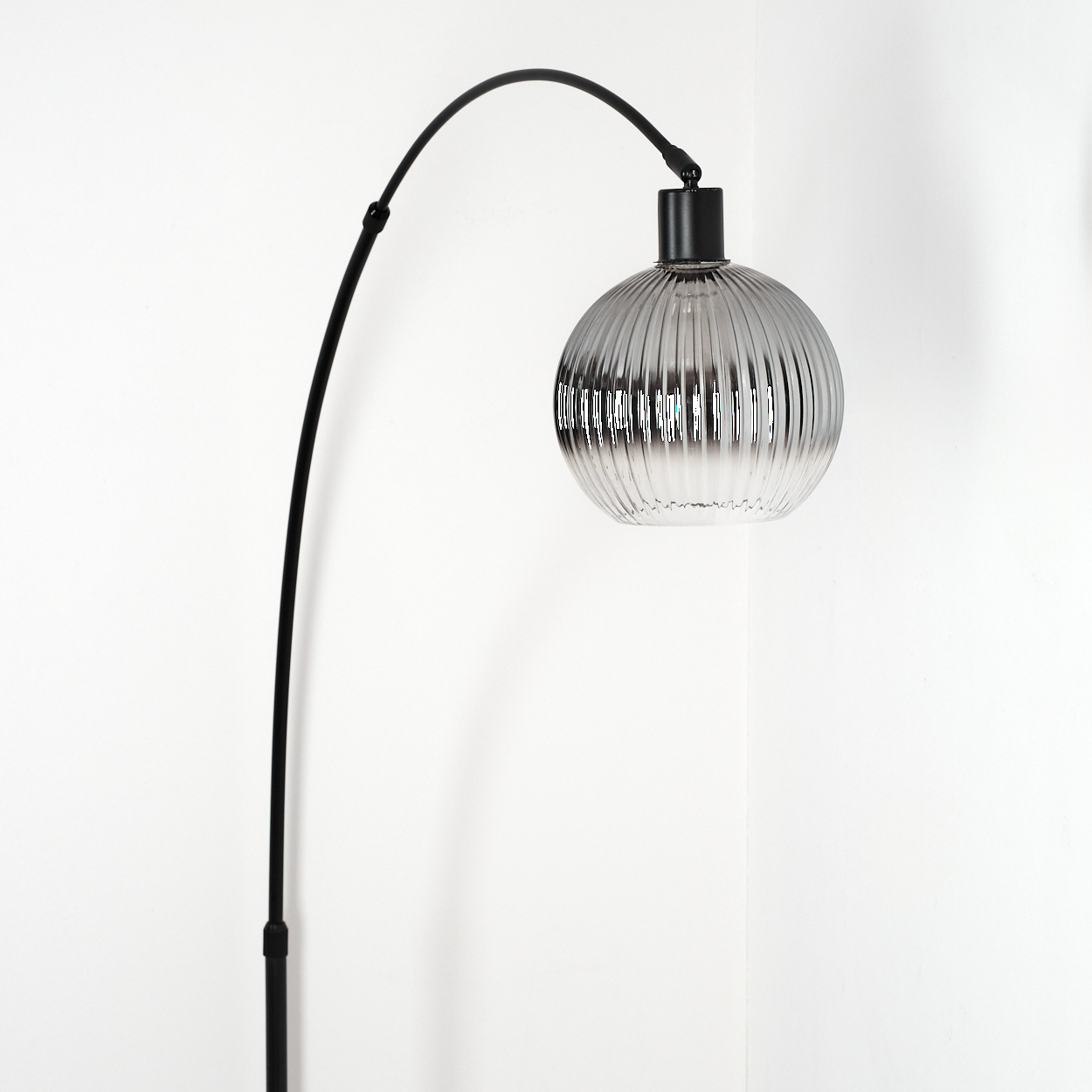 Eine moderne schwarze Bogenlampe mit geriffeltem, grauem Glasschirm. Die Lampe steht vor einer weißen Eckwand.