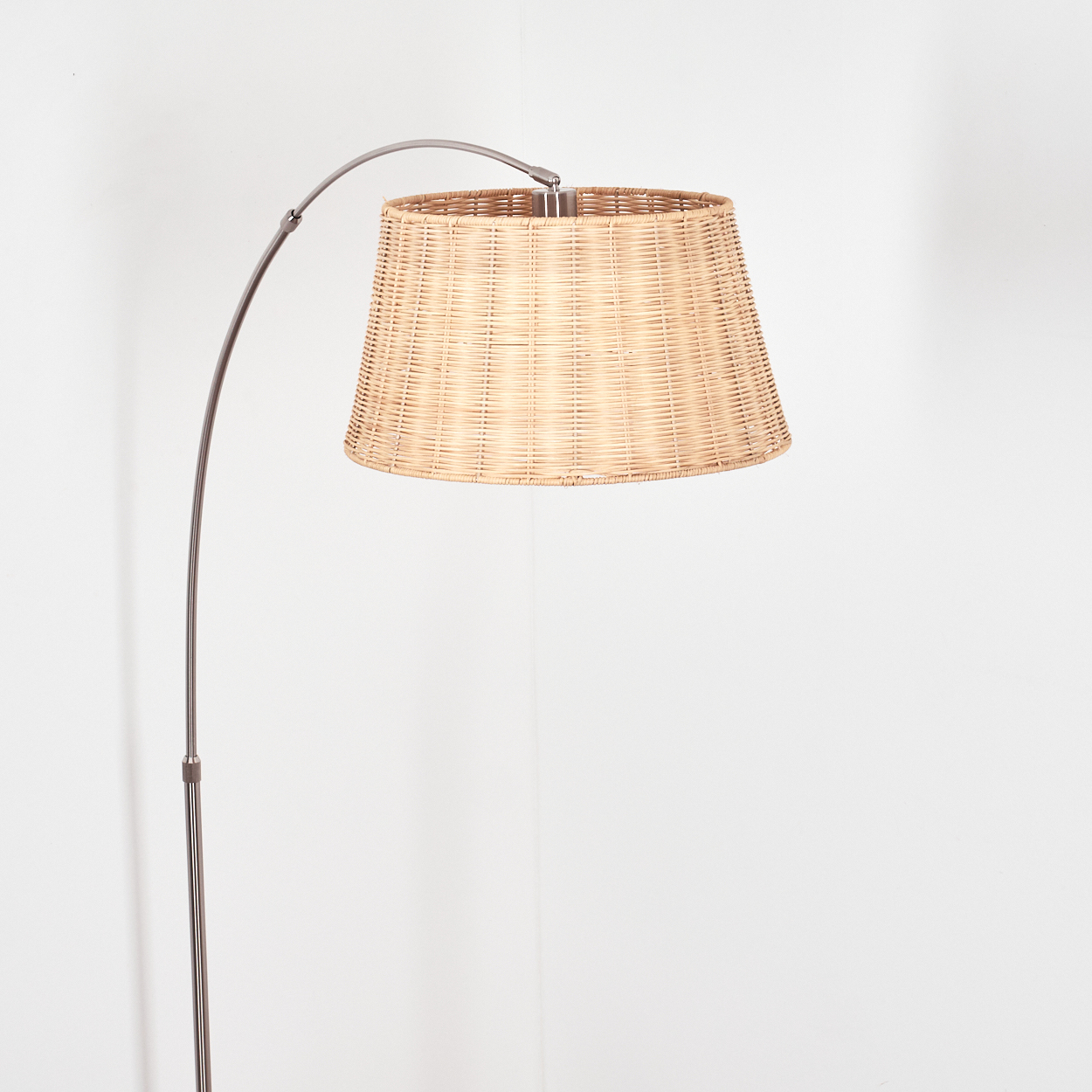 Eine Stehlampe mit gewebtem Lampenschirm. Der Metallrahmen ist gebogen, um den Lampenschirm zu stützen. Der Hintergrund ist eine weiße Wand.