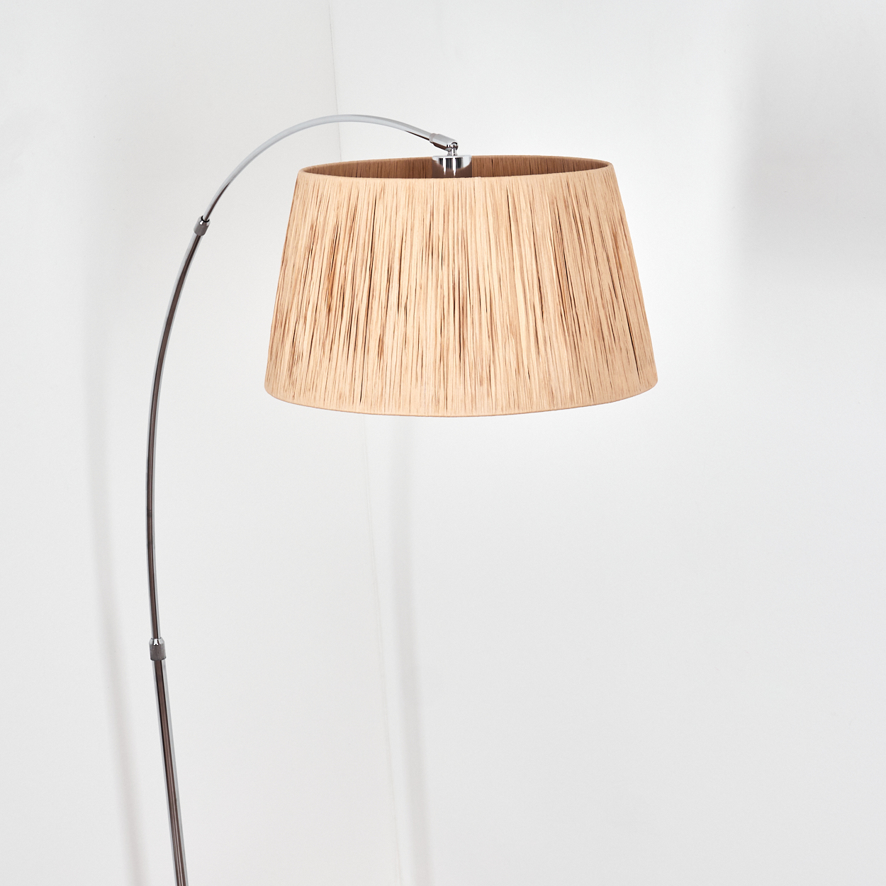 Ein beigefarbener Lampenschirm hängt an einem gebogenen Metallarm. Die Lampe steht vor einer weißen Wand. Der Schirm hat eine strukturierte Oberfläche.