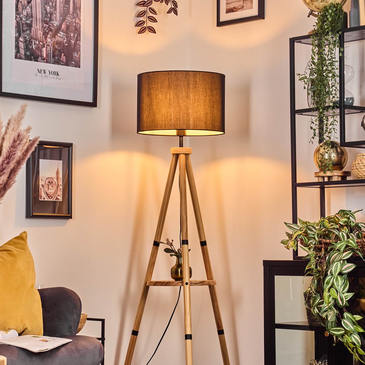Eine hölzerne Stativlampe mit schwarzem Lampenschirm. Weitere Dekorationen sind Bilderrahmen, Pflanzen und ein gelbes Kissen. Der Raum ist gut beleuchtet.