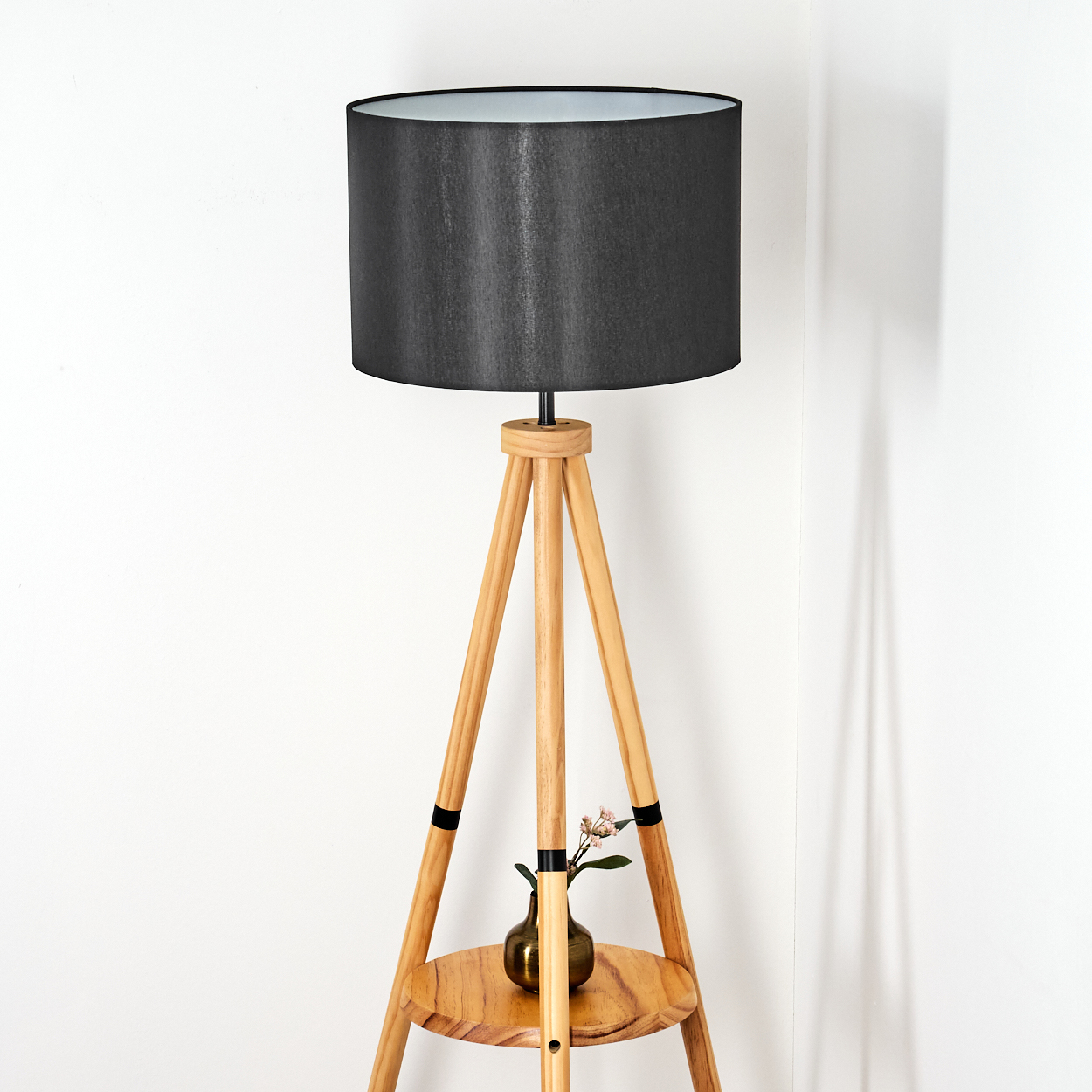Eine Stehlampe aus Holz mit schwarzem Lampenschirm. Eine kleine Vase mit Blumen steht auf einem Regal. Der Hintergrund ist eine weiße Wand.