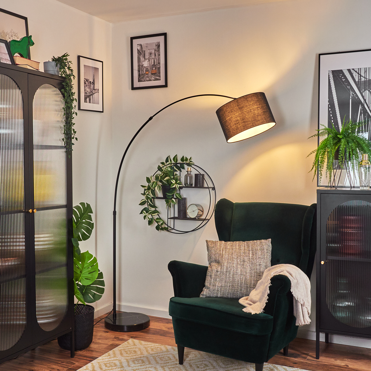Ein dunkelgrüner Sessel mit Kissen und Decke. Eine schwarze Stehlampe wölbt sich darüber. Pflanzen und gerahmte Bilder sind sichtbar.