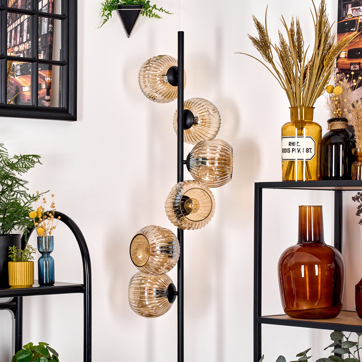 Eine moderne Lampe mit Glaskugeln steht rechts. Regale zeigen Vasen und Pflanzen, ein Bilderrahmen links.
