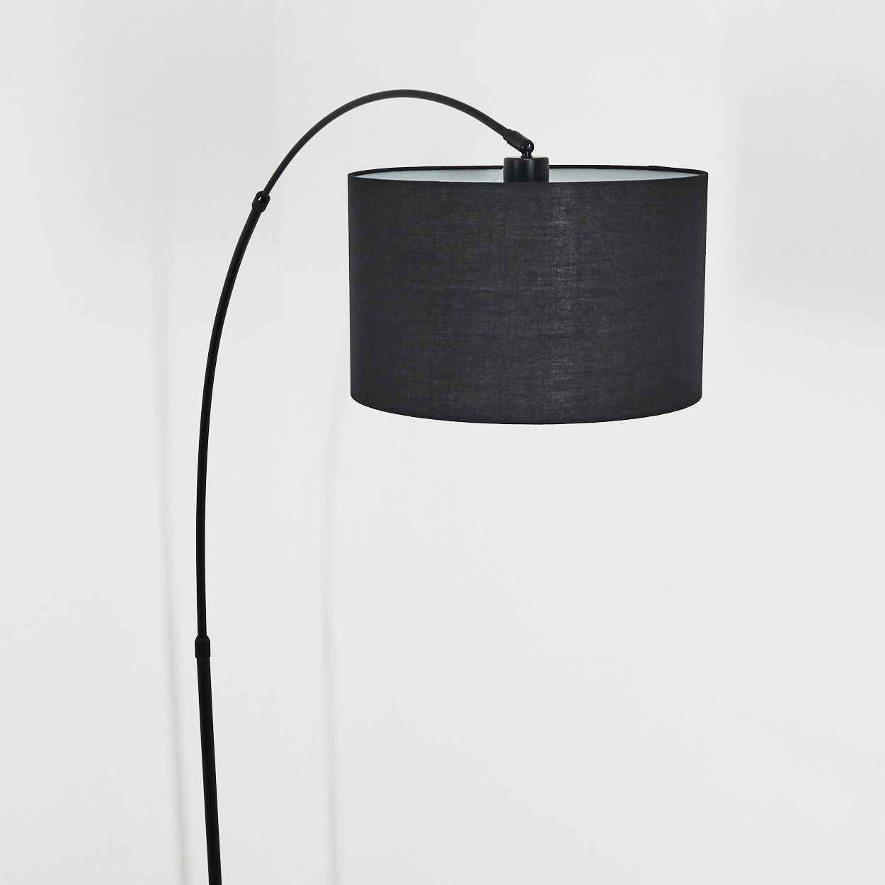 Eine schwarze, bogenförmige Stehlampe mit zylindrischem Lampenschirm. Der Hintergrund ist eine weiße Wand.