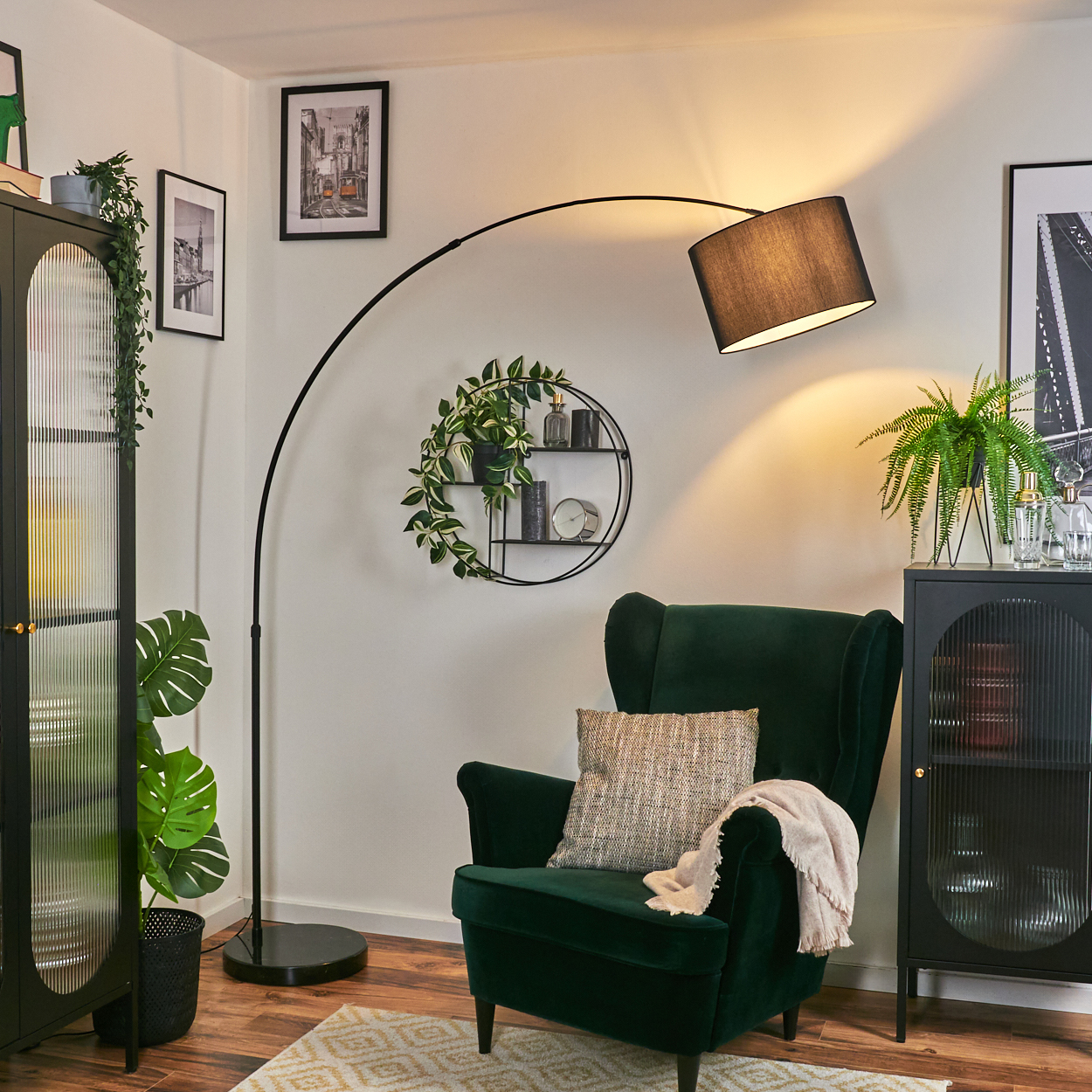 Ein Zimmer mit grünem Sessel, schwarzer Stehlampe und schwarzem Schrank. Pflanzen und Bilder schmücken die weißen Wände.