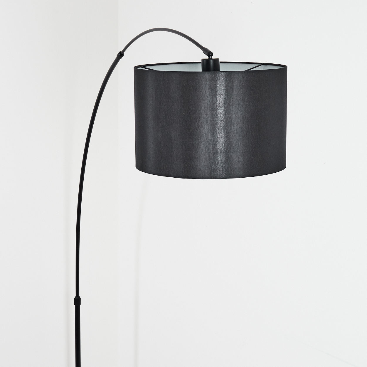 Eine moderne schwarze Stehlampe mit gebogenem Arm und zylindrischem Lampenschirm vor weißer Wand.