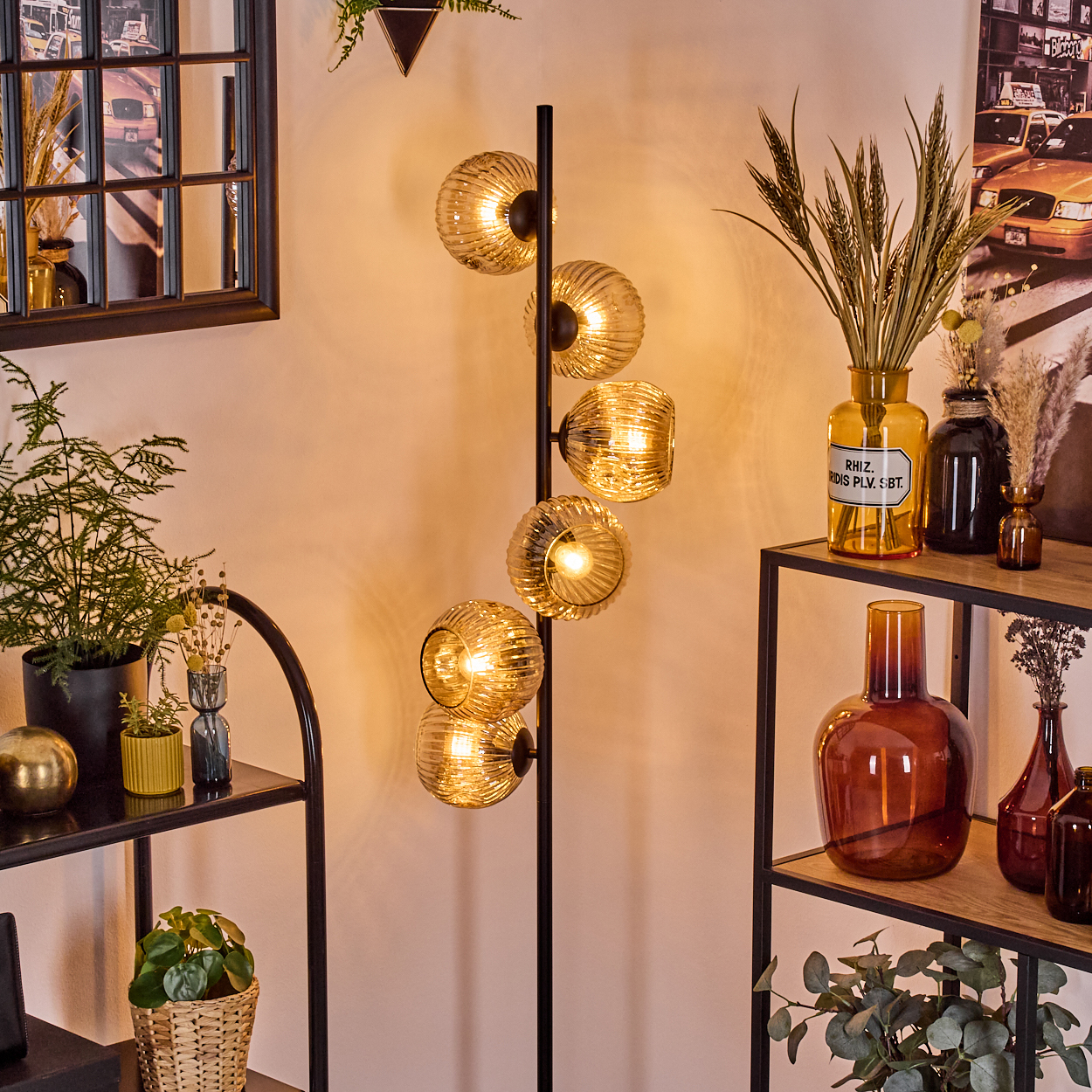 Ein modernes Interieur mit Stehlampe. Es gibt Glaskugeln, Regale mit Pflanzen und Vasen. Ein Spiegel und ein Bild sind sichtbar.
