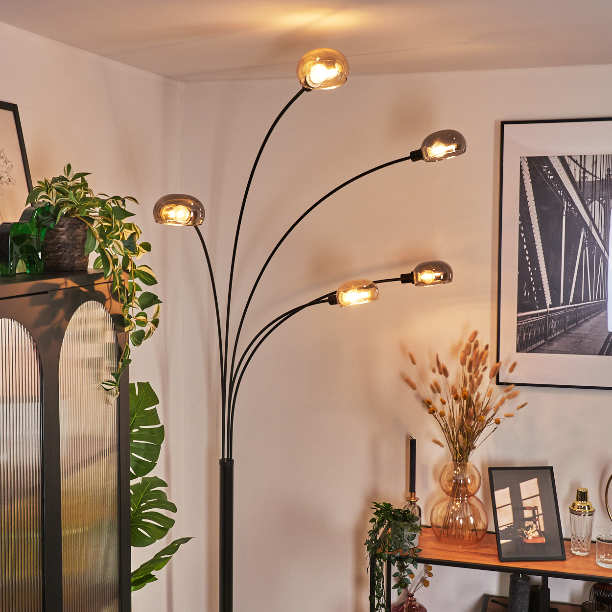 Ein modernes Wohnzimmer mit einer schwarzen Stehlampe mit fünf Glühbirnen. Pflanzen, ein Bilderrahmen und ein Holzregal sind ebenfalls zu sehen.