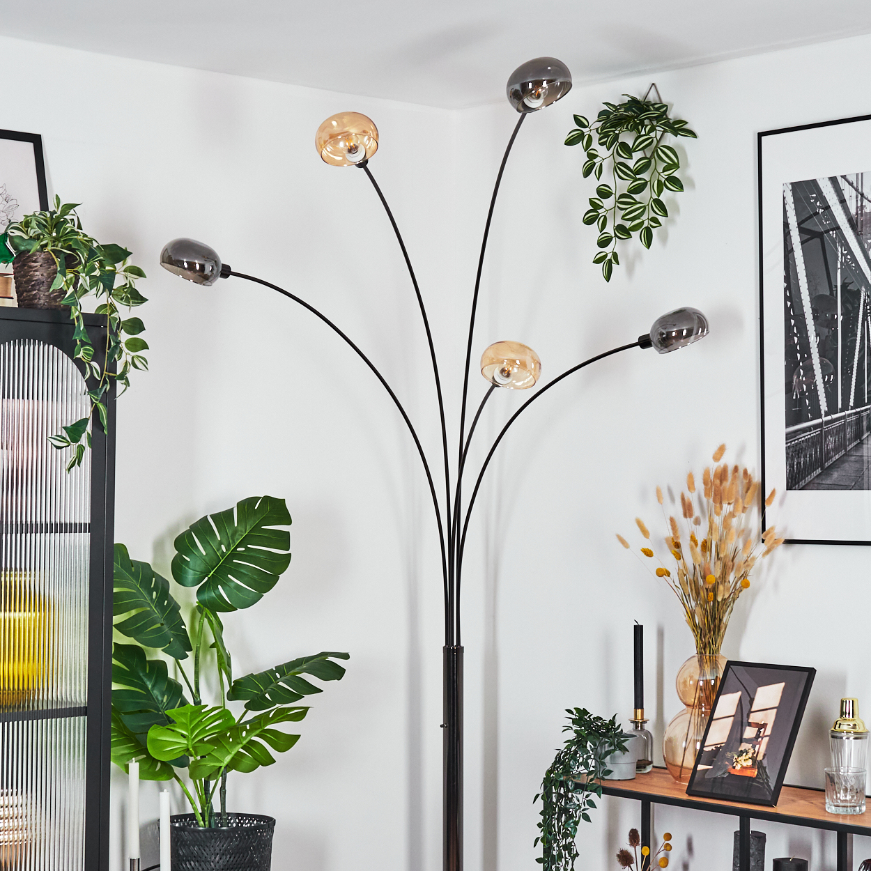 Ein modernes Wohnzimmer mit schwarzer Lampe, Pflanzen und einem gerahmten Bild. Die Lampe hat fünf Lichter. Die Wände sind weiß.