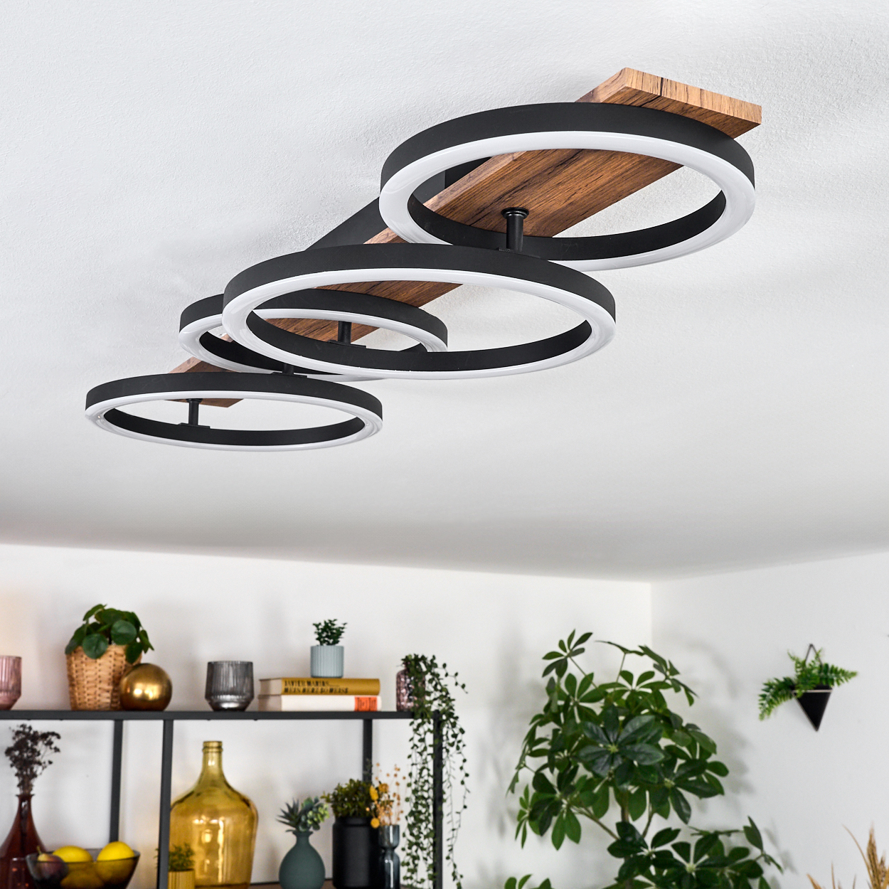 Eine moderne Deckenleuchte mit schwarzen Ringen und weißen Innenlichtern auf Holzbasis. Pflanzen und Regale im Hintergrund.