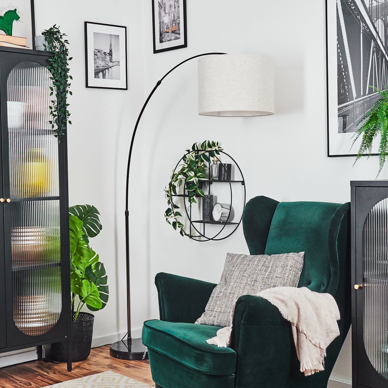 Ein grüner Sessel mit Kissen und Decke, eine Stehlampe, ein schwarzer Schrank und gerahmte Bilder an einer weißen Wand.