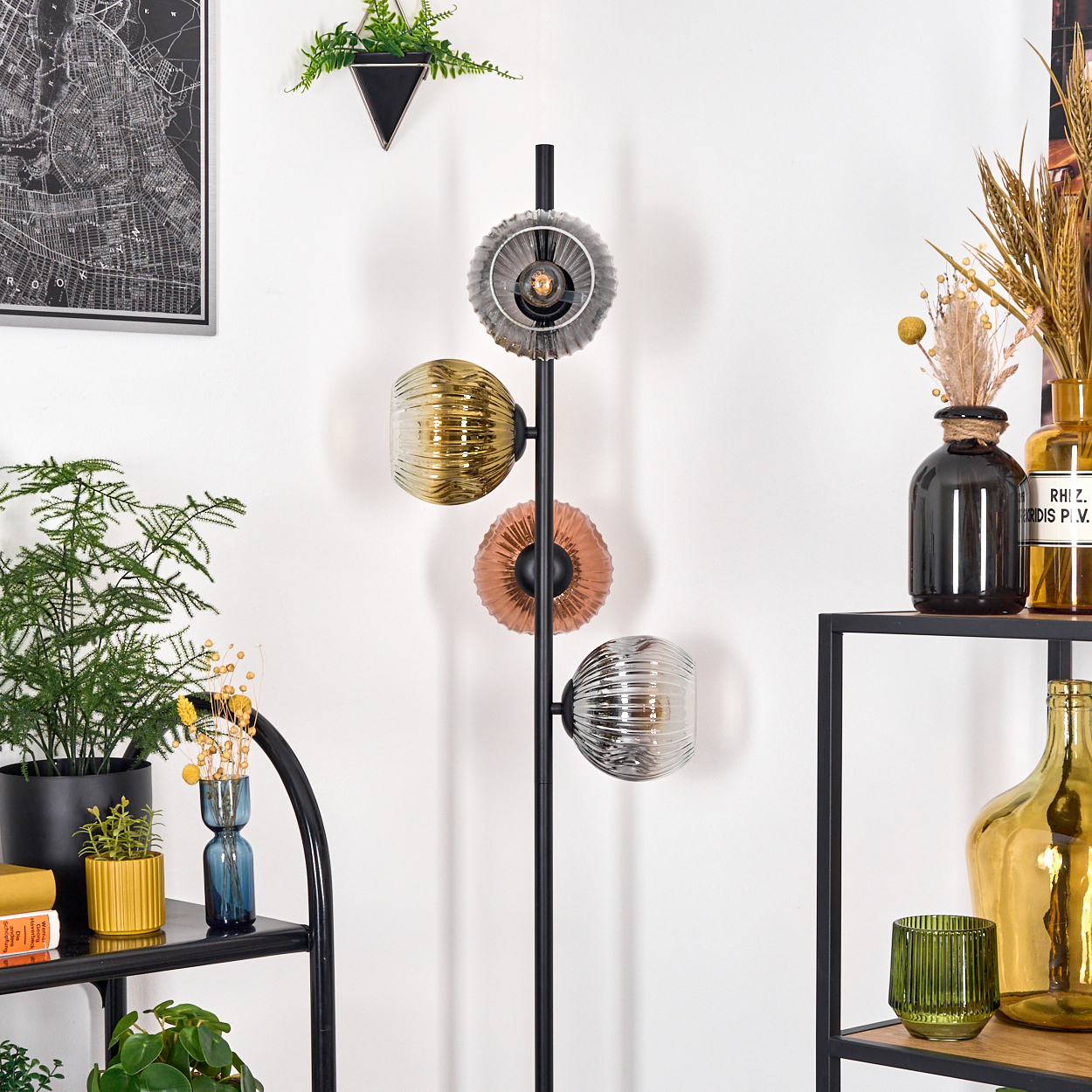 Eine moderne Stehlampe mit vier bunten Kugellampen steht vor einer weißen Wand, Pflanzen und Regale sind sichtbar.