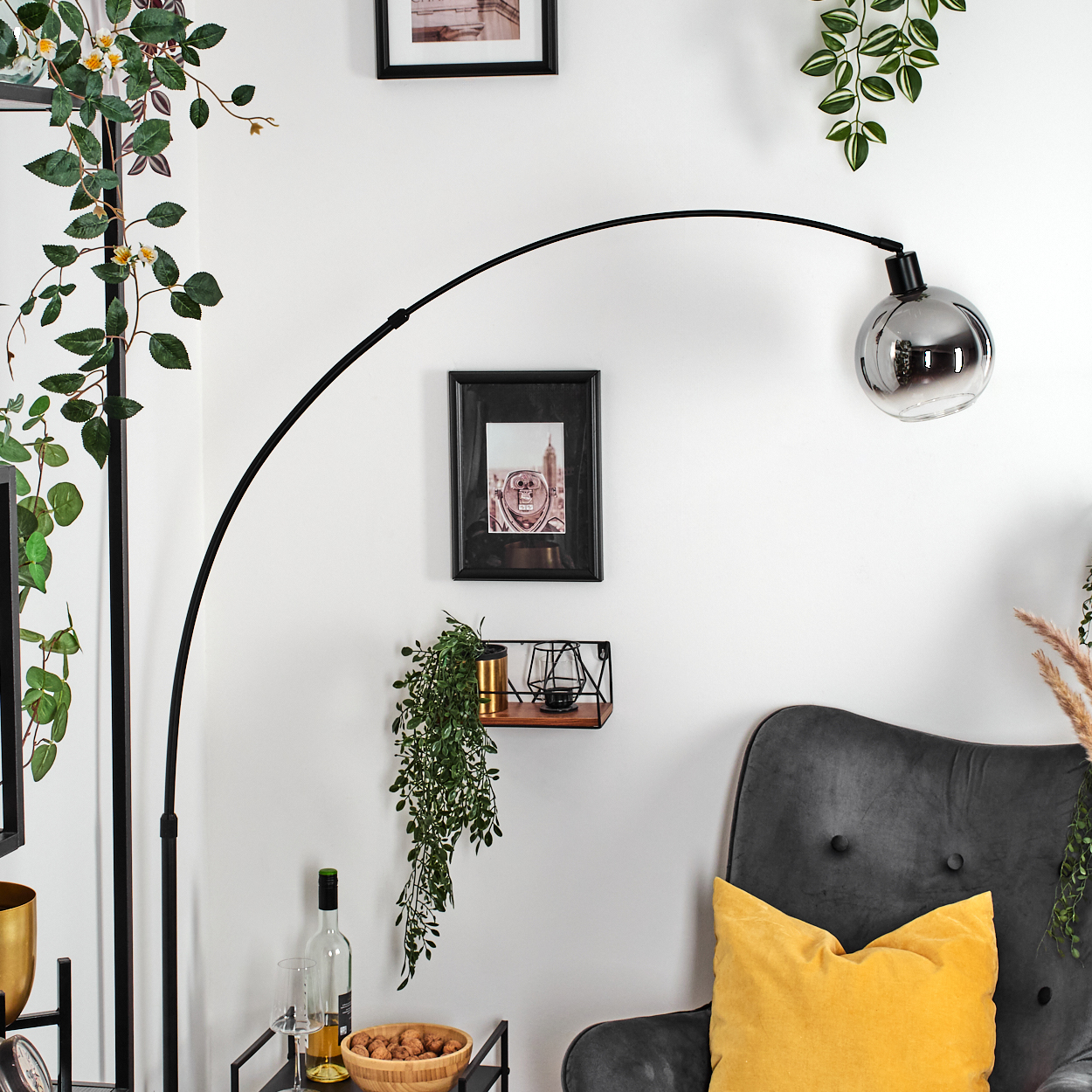 Ein Raum mit einer schwarzen Bogenlampe, einem grauen Sessel mit gelbem Kissen und einem Tisch mit einer Weinflasche.