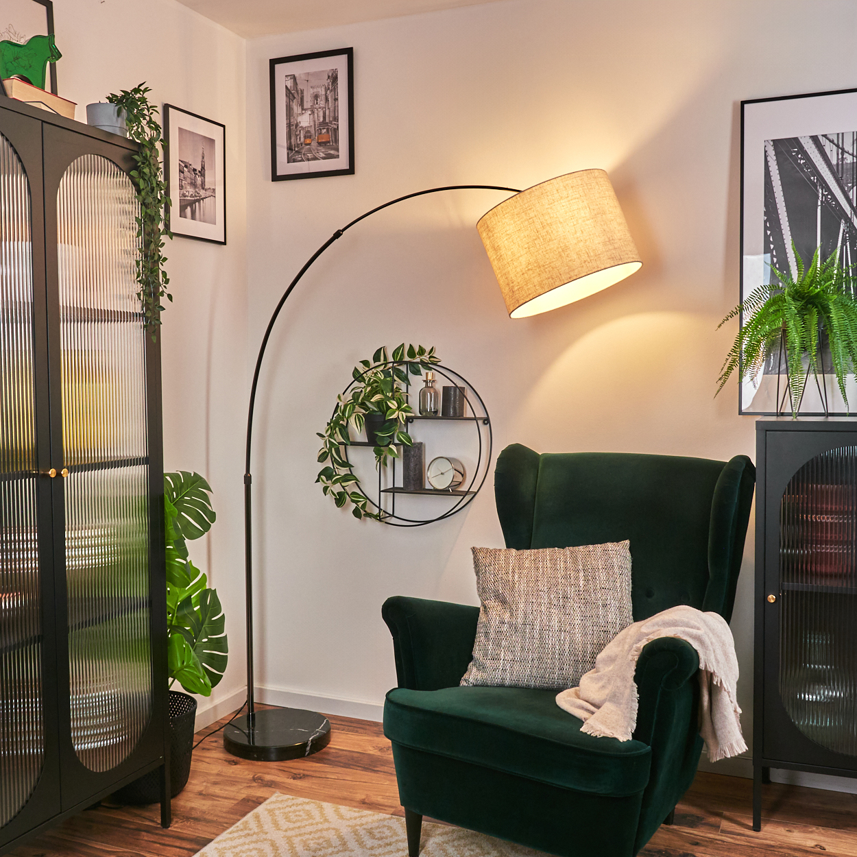 Ein grüner Sessel mit Kissen und Decke steht unter einer Lampe. Schwarze Schränke und Pflanzen sind im Hintergrund. Bilder hängen an den Wänden.