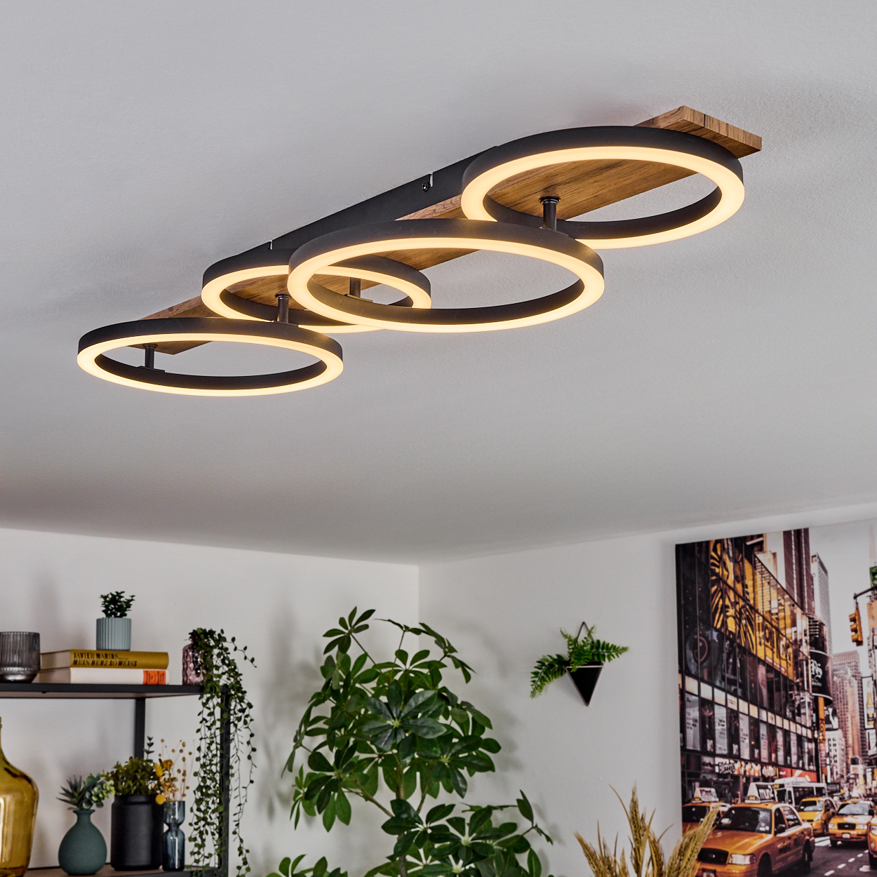 Eine moderne Deckenleuchte mit vier beleuchteten Ringen. Darunter Pflanzen und ein Regal mit Büchern. Ein New York City Bild an der Wand.