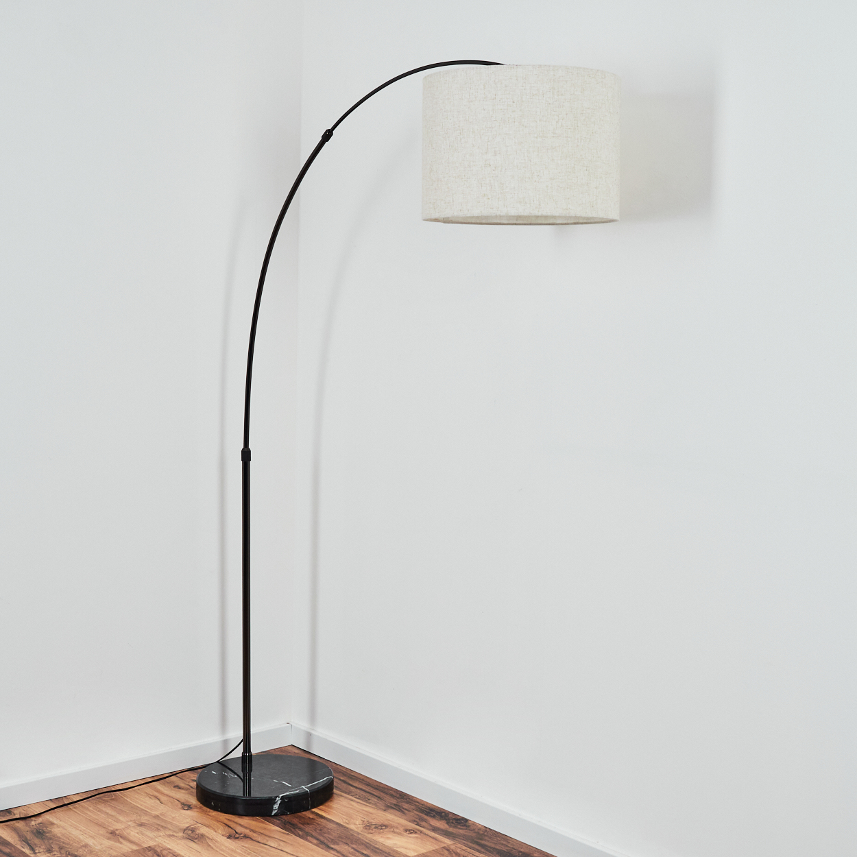 Eine schwarze, bogenförmige Stehlampe mit beigem Lampenschirm steht in einer Zimmerecke. Die Wände sind weiß, der Boden ist aus Holz.