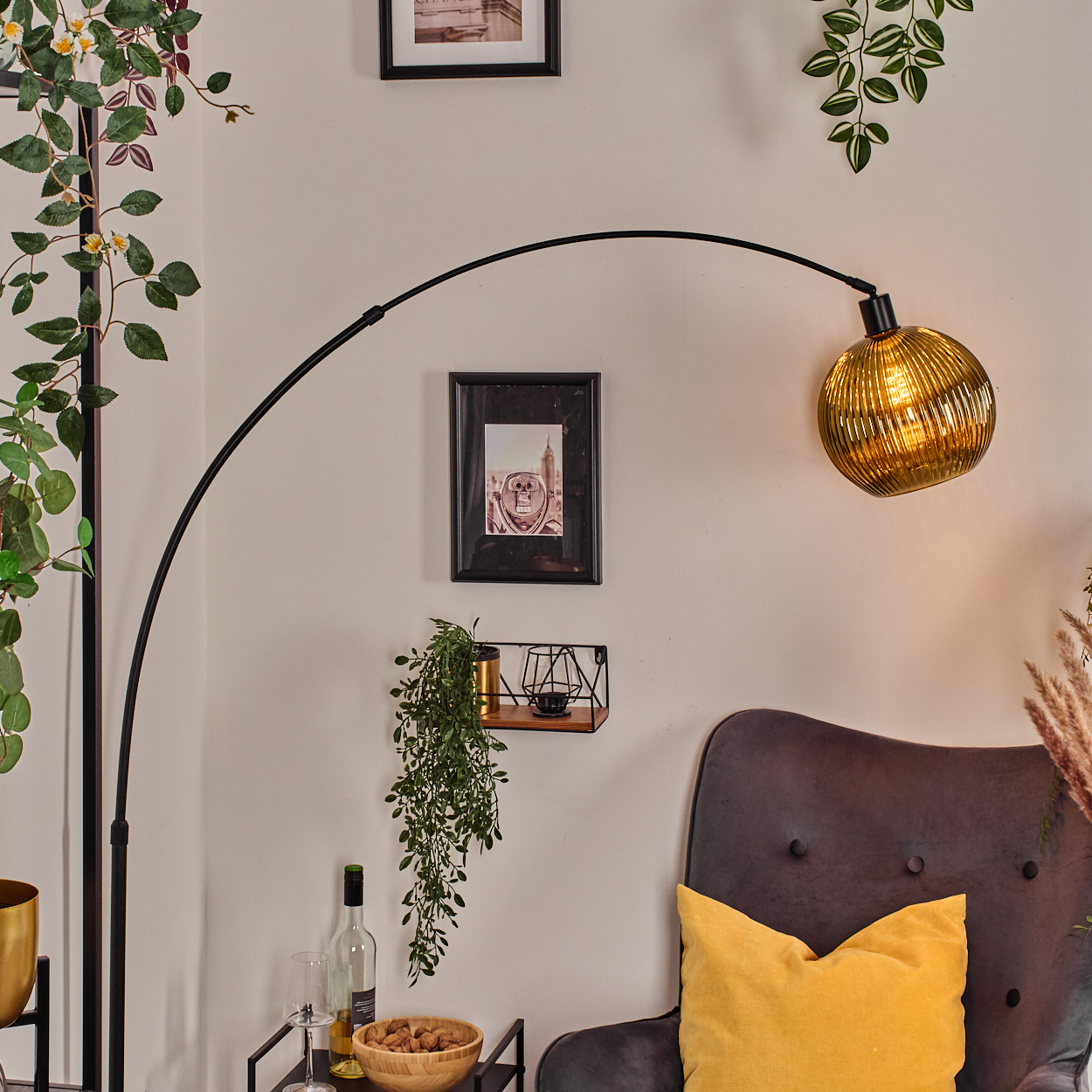 Ein Raum mit einer schwarzen Bogenlampe, einem dunkelgrauen Sessel, einem gelben Kissen und einer goldenen Lampe. Bilder und Pflanzen an der Wand.