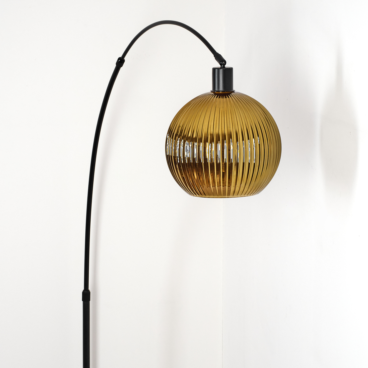Eine moderne Bogenstehlampe mit kugelförmigem, geripptem bernsteinfarbenem Glasschirm vor einer weißen Ecke.