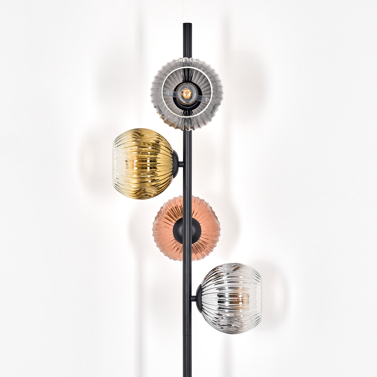 Eine moderne Wandlampe mit vier Glasschirmen in Silber, Gold, Kupfer und Silber. Ein schwarzer Metallstab hält die Schirme an einer weißen Wand.