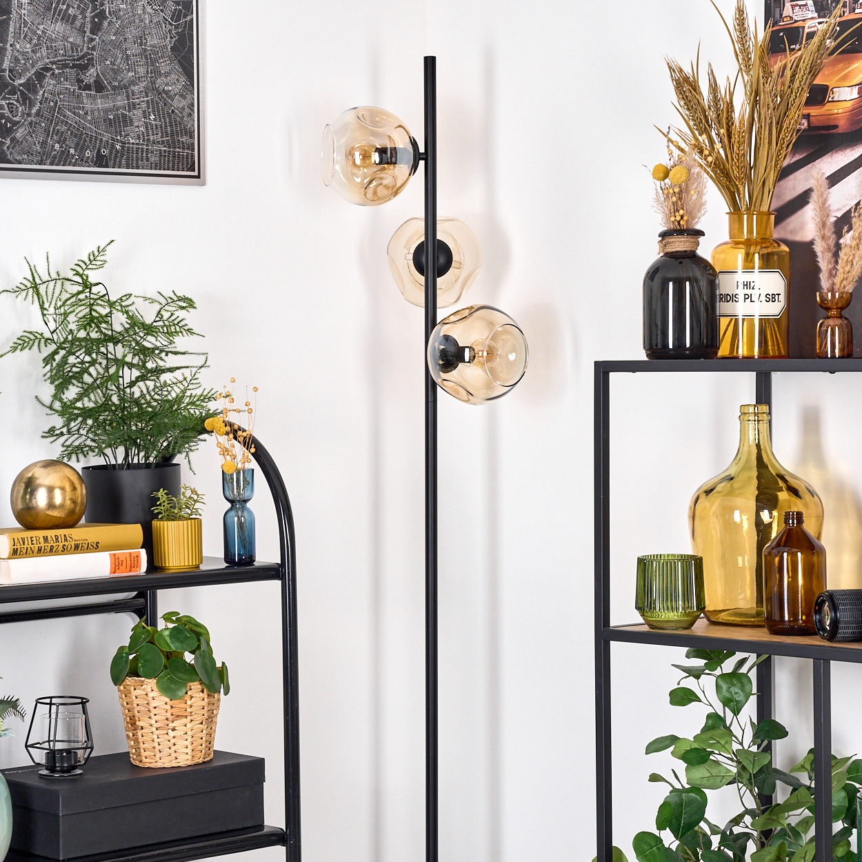 Ein Raum mit einem schwarzen Metallregal und einer Stehlampe mit drei Glasschirmen. Pflanzen und Dekorationsartikel füllen die Regale.