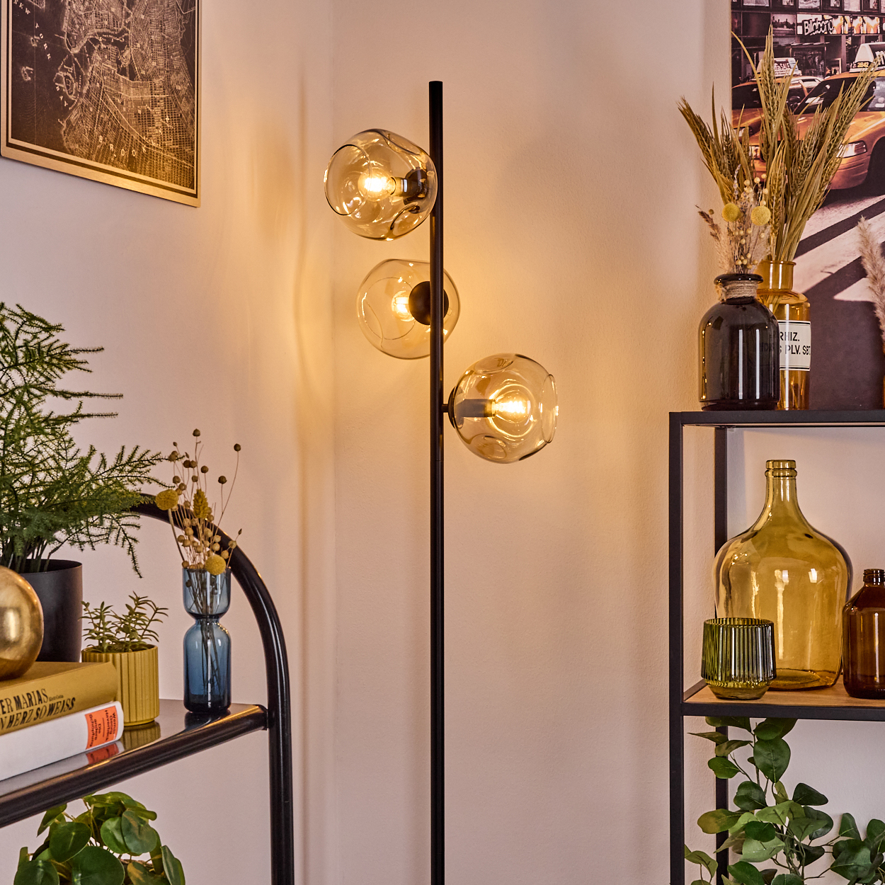 Eine moderne Lampe mit drei kugelförmigen Schirmen. Regale mit Vasen, Pflanzen und Büchern befinden sich im Vordergrund. Eine Karte und ein Stadtposter.