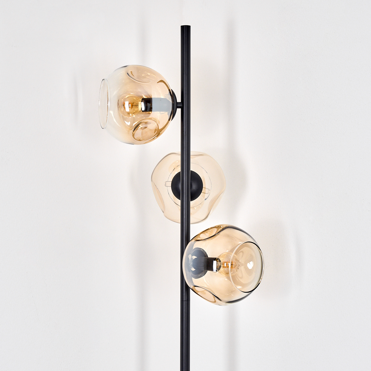 Eine moderne Stehlampe mit drei bernsteinfarbenen Glasschirmen. Die Lampe hat eine schwarze Stange und steht vor einer weißen Wand.