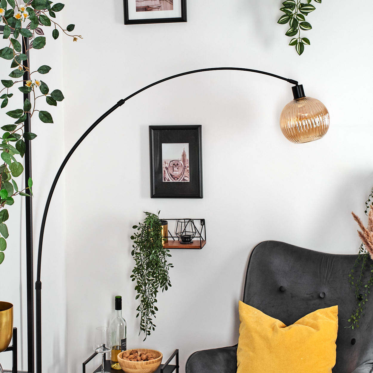 Ein modernes Wohnzimmer mit schwarzer Stehlampe, grauem Sessel mit gelbem Kissen, Pflanzen, Bildern, Wein und einer Holzschale.