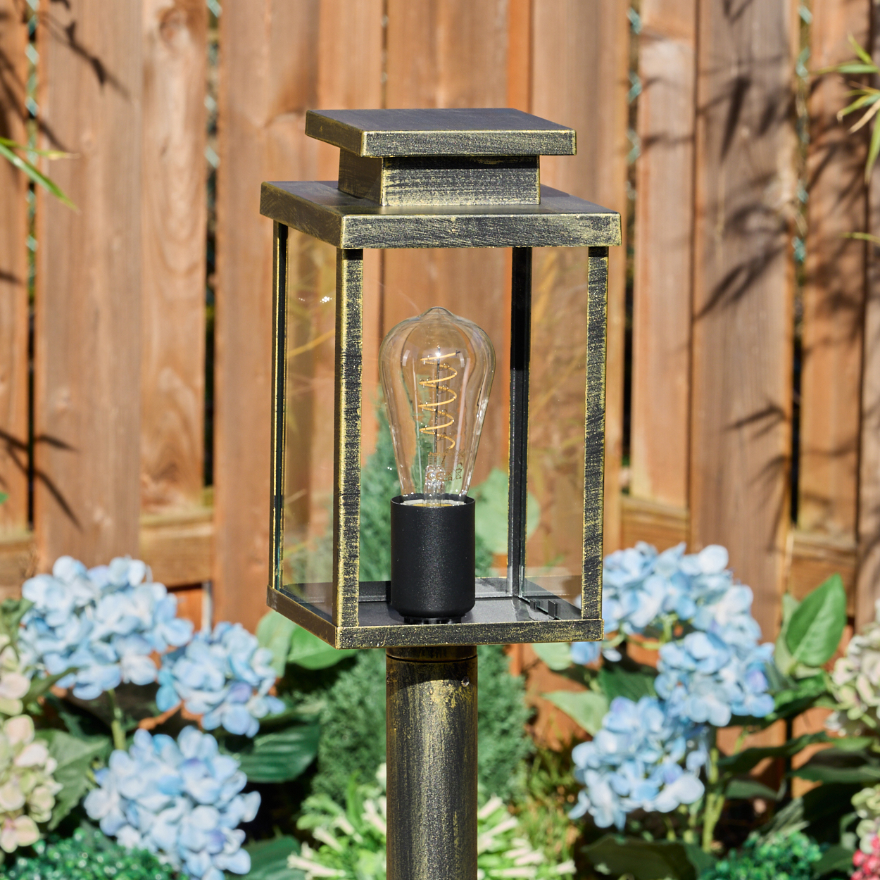 Eine dekorative Außenlampe mit einer Vintage-Glühbirne, umgeben von blauen Blumen und einem Holzzaun im Hintergrund.