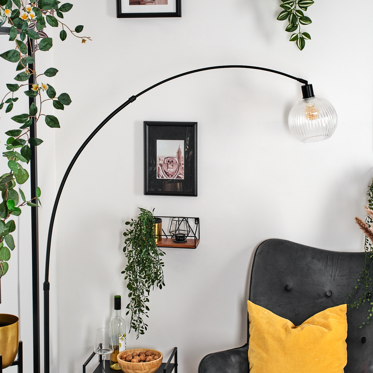 Wohnzimmerszene mit schwarzer Bogenlampe, grauem Sessel, gelbem Kissen und gerahmten Bildern an einer weißen Wand mit Pflanzen.