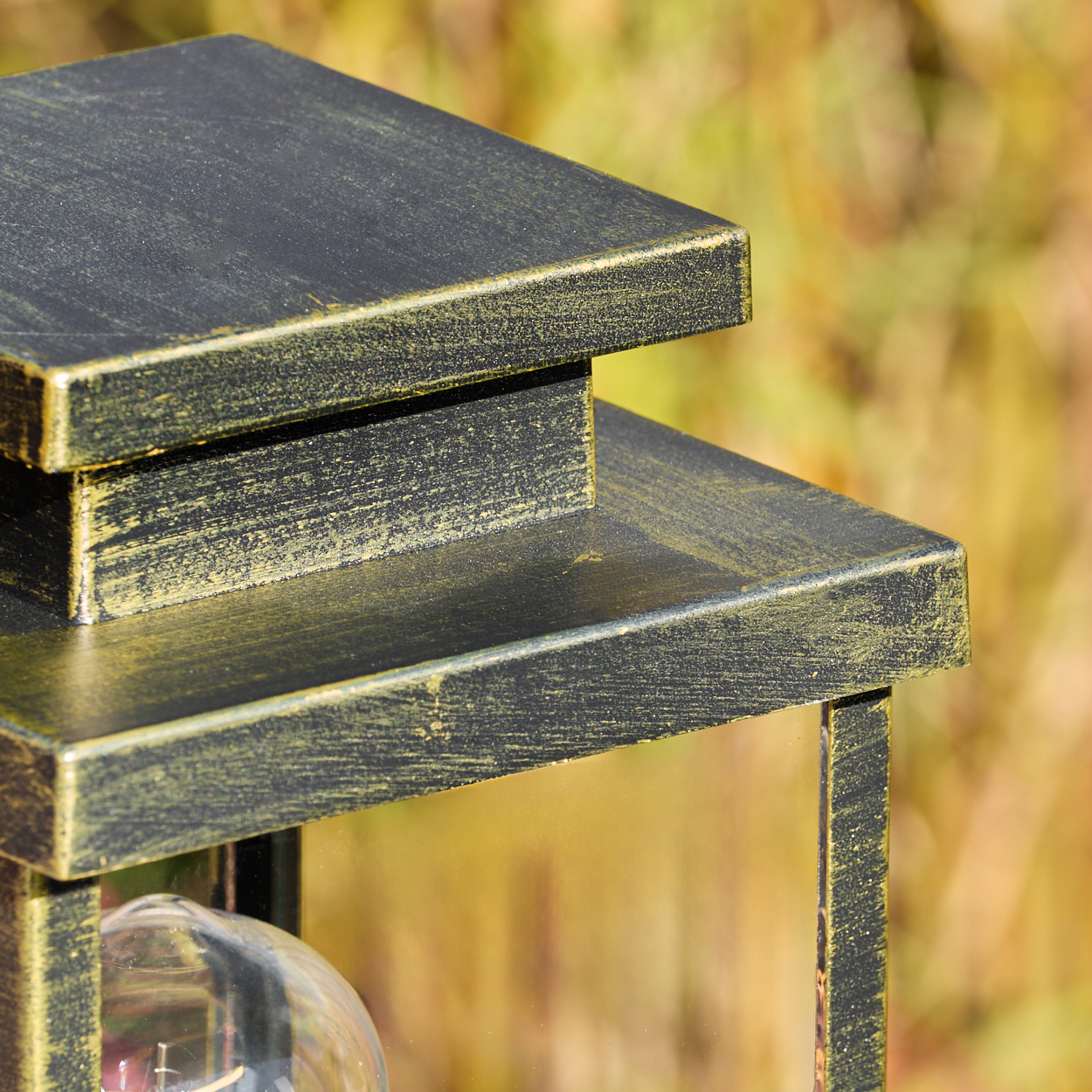 Nahaufnahme einer schwarz-goldenen Außenlampe. Die Lampe hat ein mehrschichtiges Design mit sichtbarer Glühbirne. Der Hintergrund ist verschwommen grün.