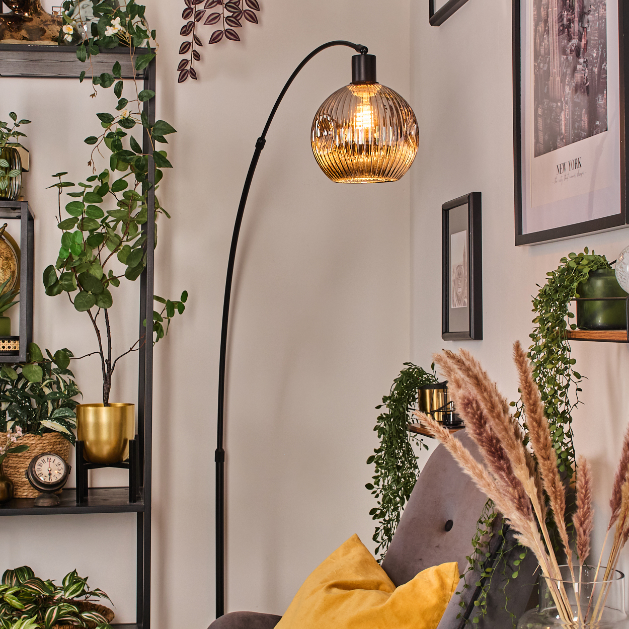 Ein Raum mit einer schwarzen Bogenlampe, Pflanzen, einem goldenen Topf, einem grauen Stuhl mit gelbem Kissen und Bildern an der Wand.