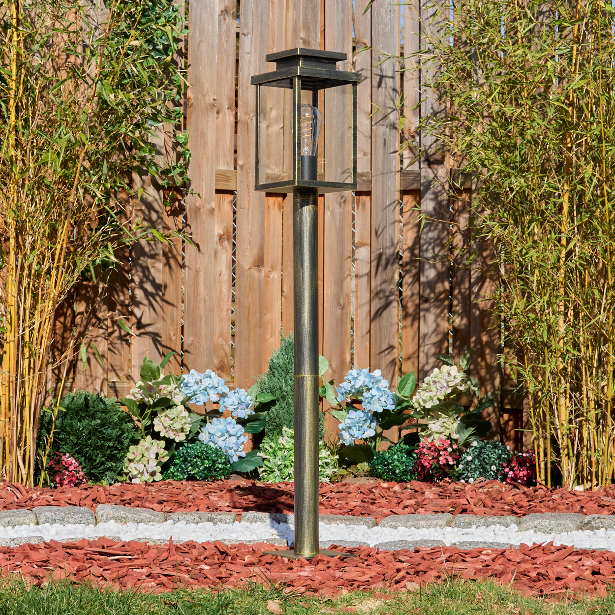 Eine Außenlampe in einem Garten mit Holzzaun im Hintergrund, umgeben von Pflanzen und Blumen, mit grünem Gras im Vordergrund.