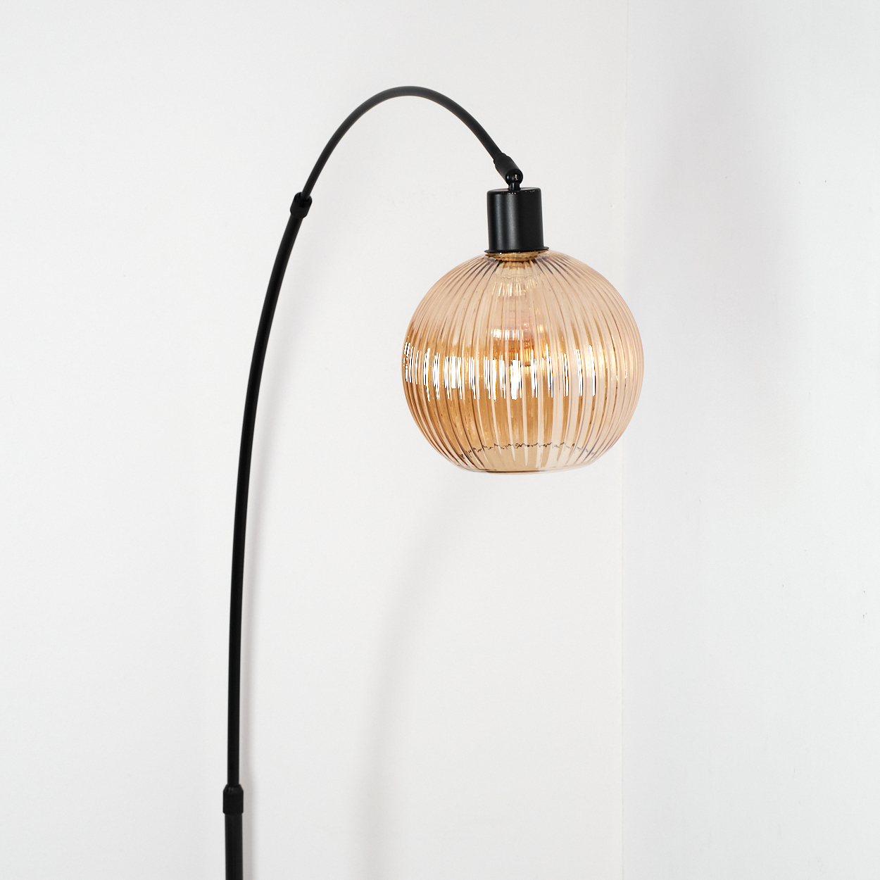 Eine moderne Stehlampe mit schwarzem gebogenem Arm und rundem, geripptem, bernsteinfarbenem Glasschirm vor weißer Wand.