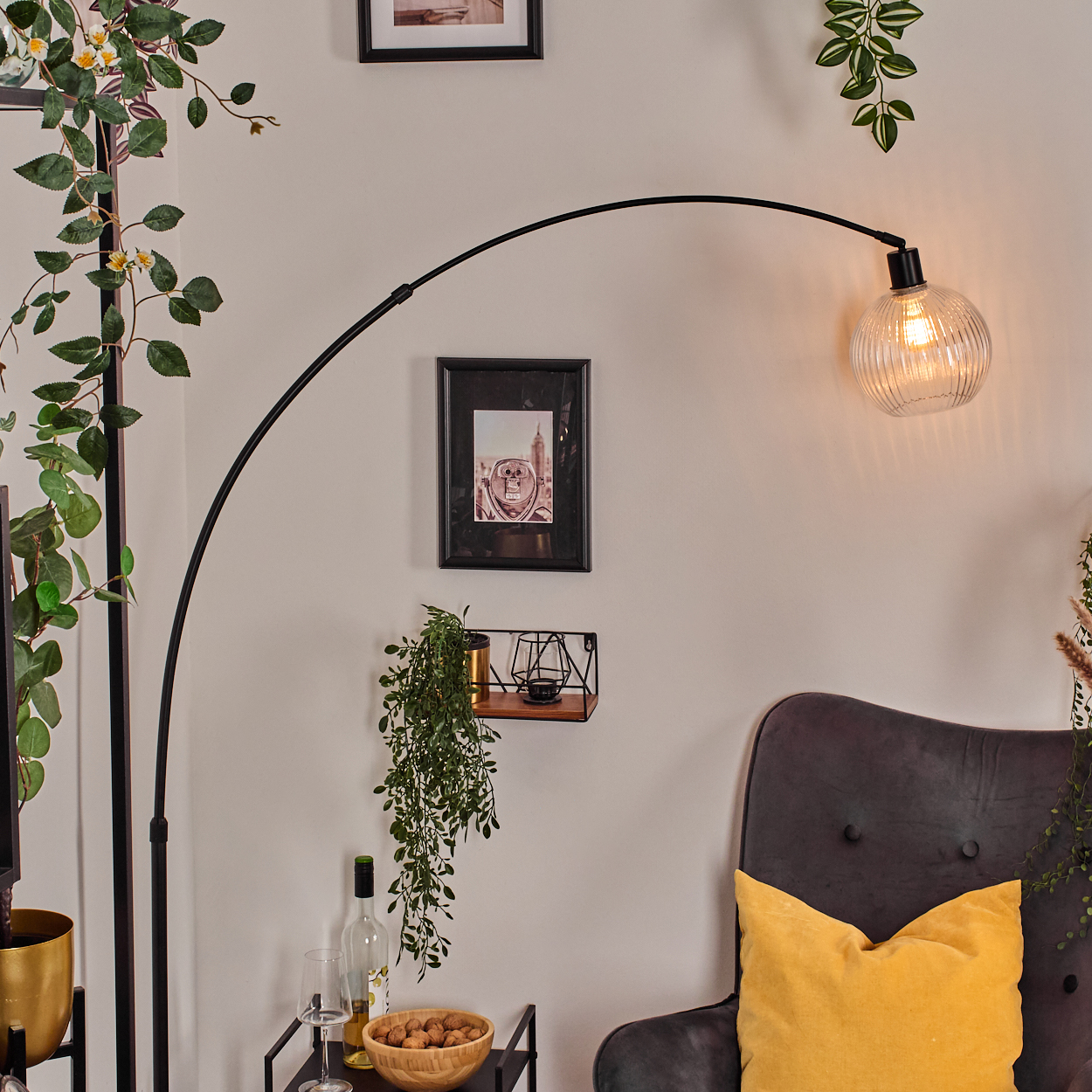 Ein Raum mit einer schwarzen Lampe, einem Stuhl mit gelbem Kissen und einem kleinen Tisch mit Flasche, Glas und Walnüssen. Überall Pflanzen.