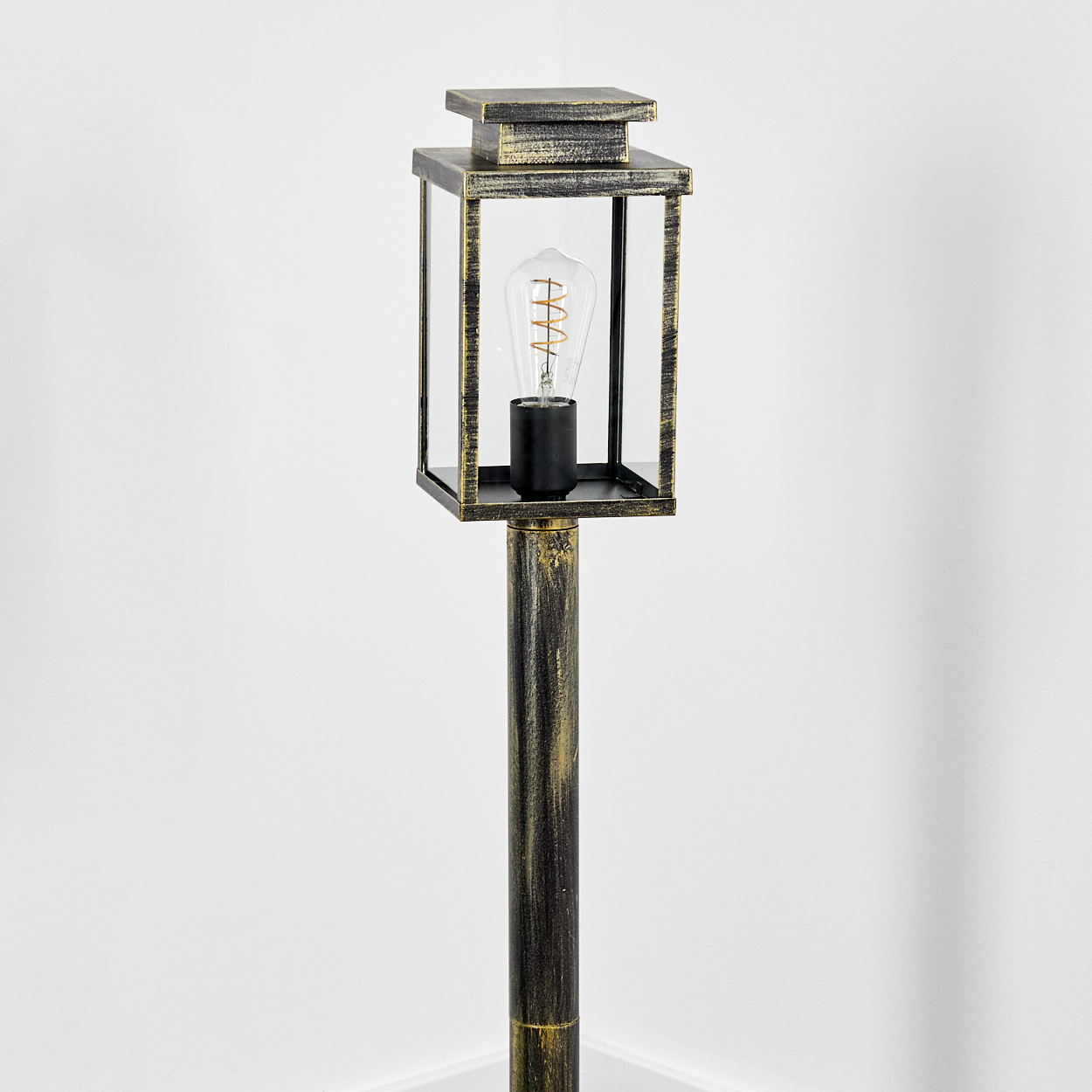 Eine Außenlaterne mit quadratischer Leuchte, Glasscheiben, einer dekorativen Glühbirne und dunklem Metallfinish. Vor einer weißen Wand.