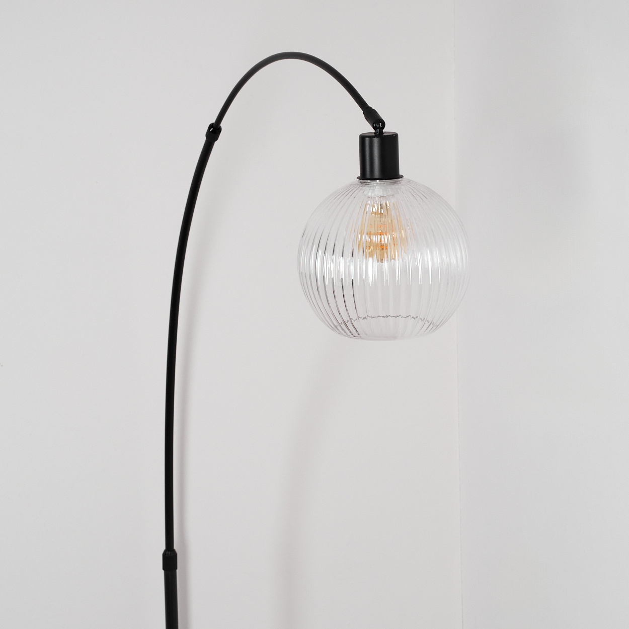 Eine moderne schwarze Bogenlampe mit klarem, geripptem Globus-Schirm vor einer weißen Wand.