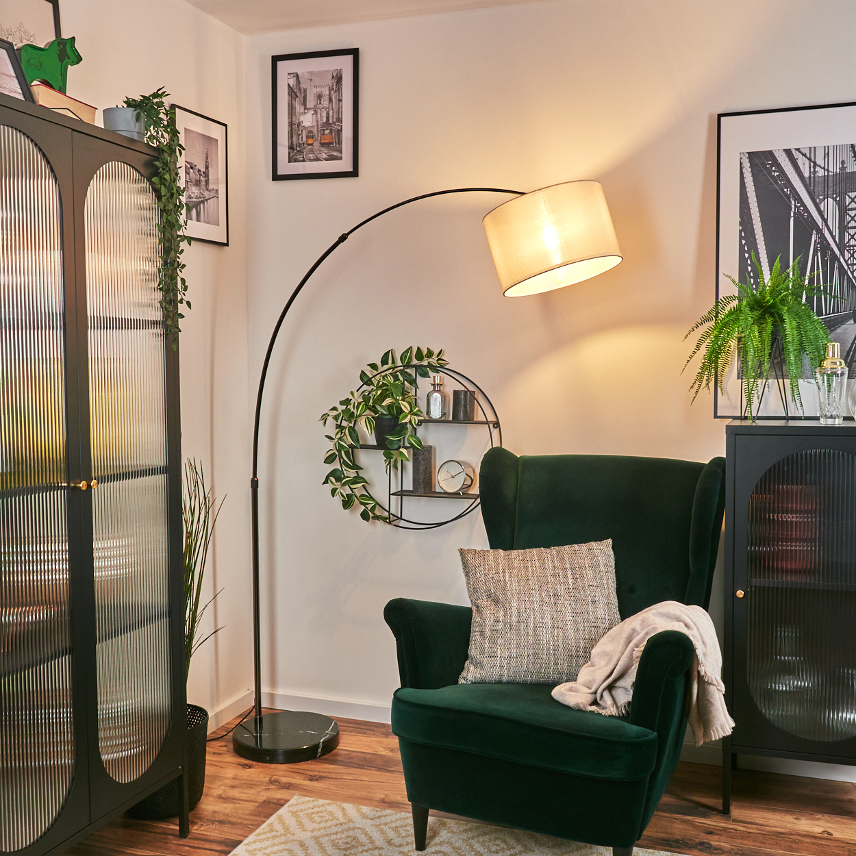 Ein grüner Sessel mit Kissen und Decke. Eine Stehlampe beleuchtet die Szene. Es gibt einen schwarzen Schrank und ein dekoratives Regal.