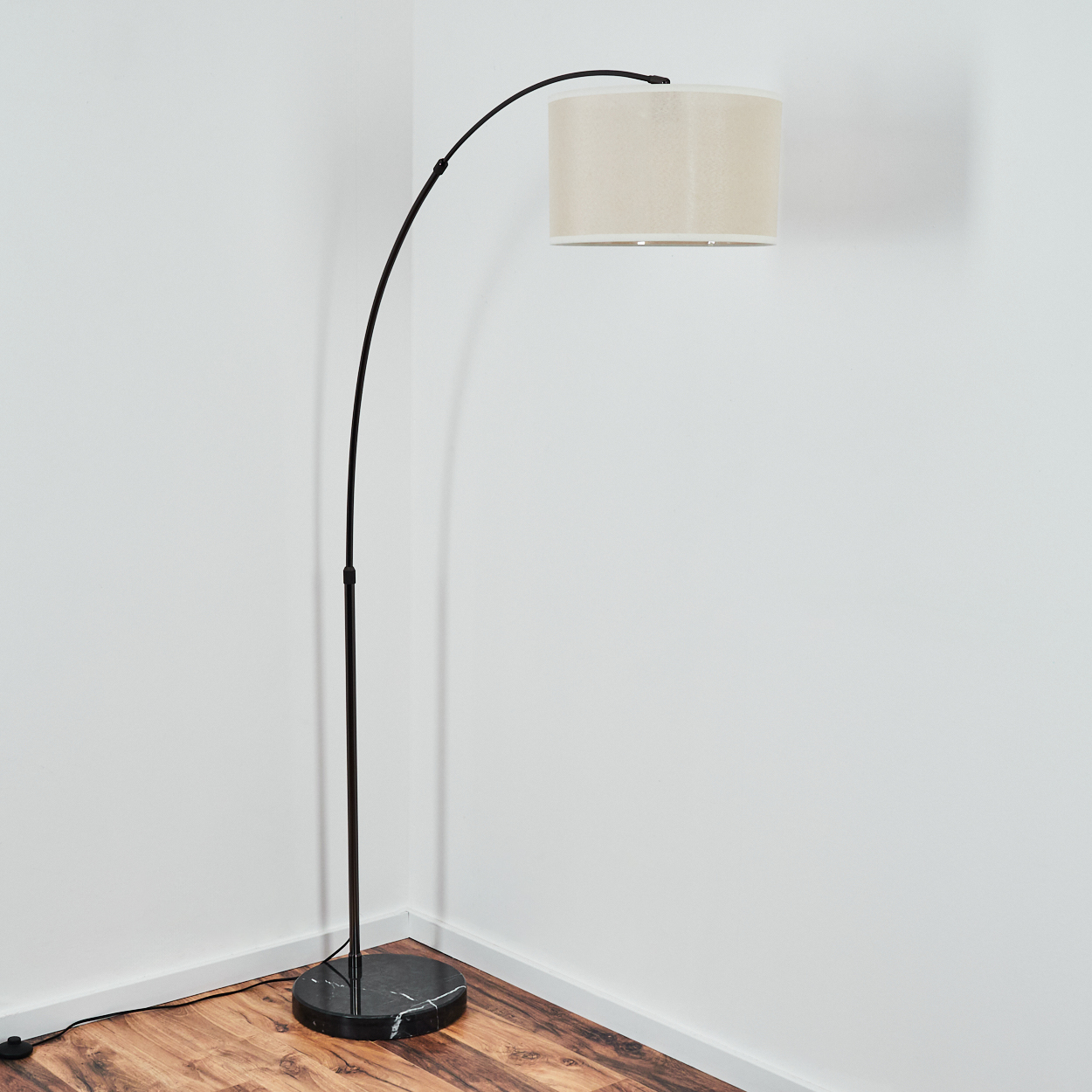 Eine moderne, schwarze Bogen-Stehlampe mit beigem Lampenschirm steht in einer Ecke vor weißen Wänden und Holzboden.