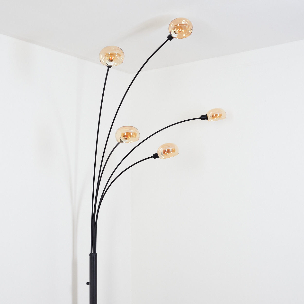 Eine moderne Stehlampe mit fünf Glühbirnen. Die Glühbirnen sind rund und bernsteinfarben, das Gestell ist schwarz. Vor einer weißen Wand.