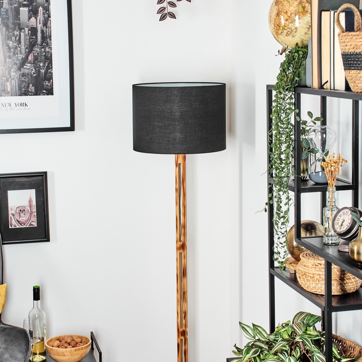 Raumecke mit Stehlampe. Schwarzer Lampenschirm, Holzständer. Regale mit Büchern, Globus, Pflanzen und Dekorationsartikeln. Wandkunst.