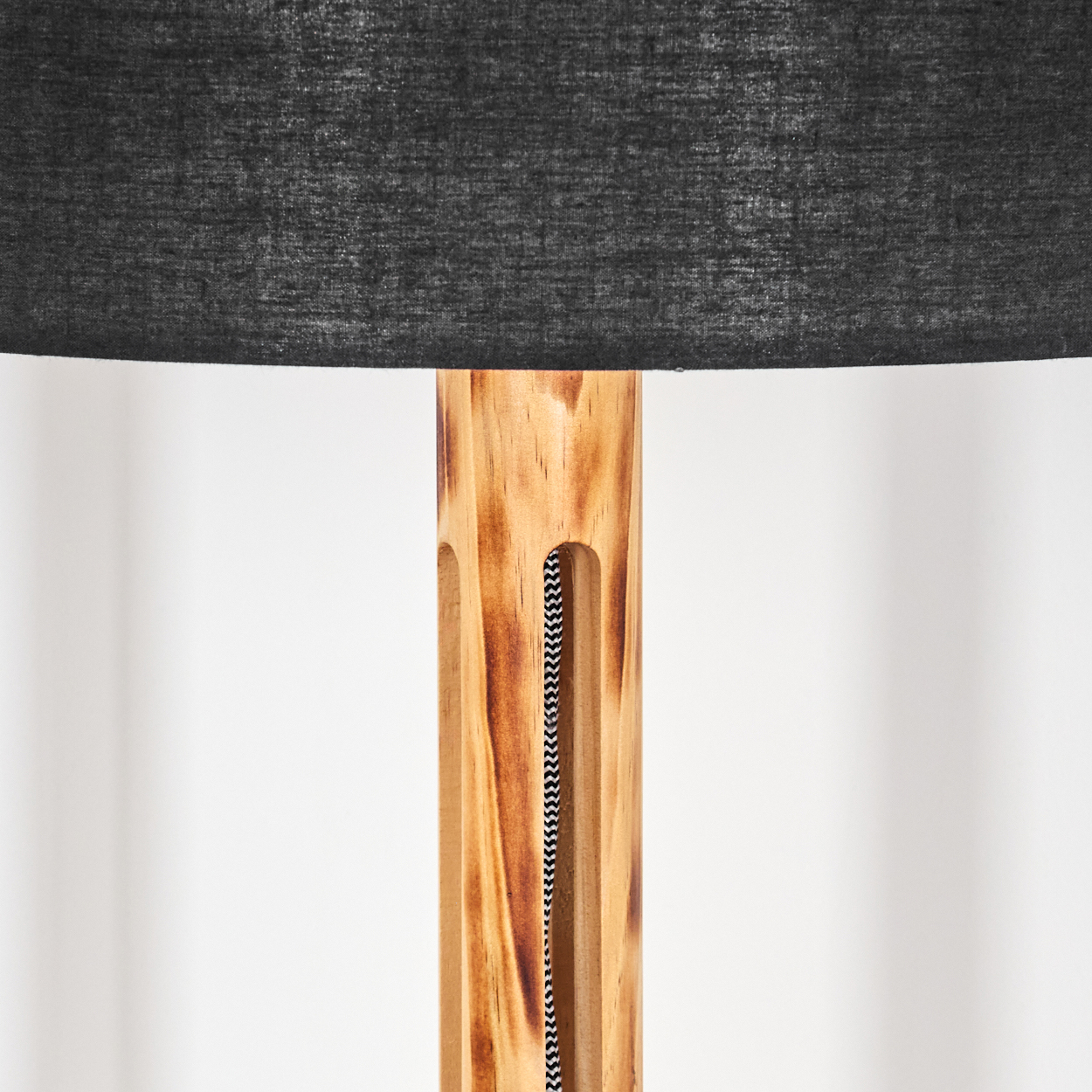 Holzlampe mit schwarzem Lampenschirm. Der Holzfuß hat ein schwarz-weiß gestreiftes Kabel, das durch ihn hindurchführt, vor einer weißen Wand.