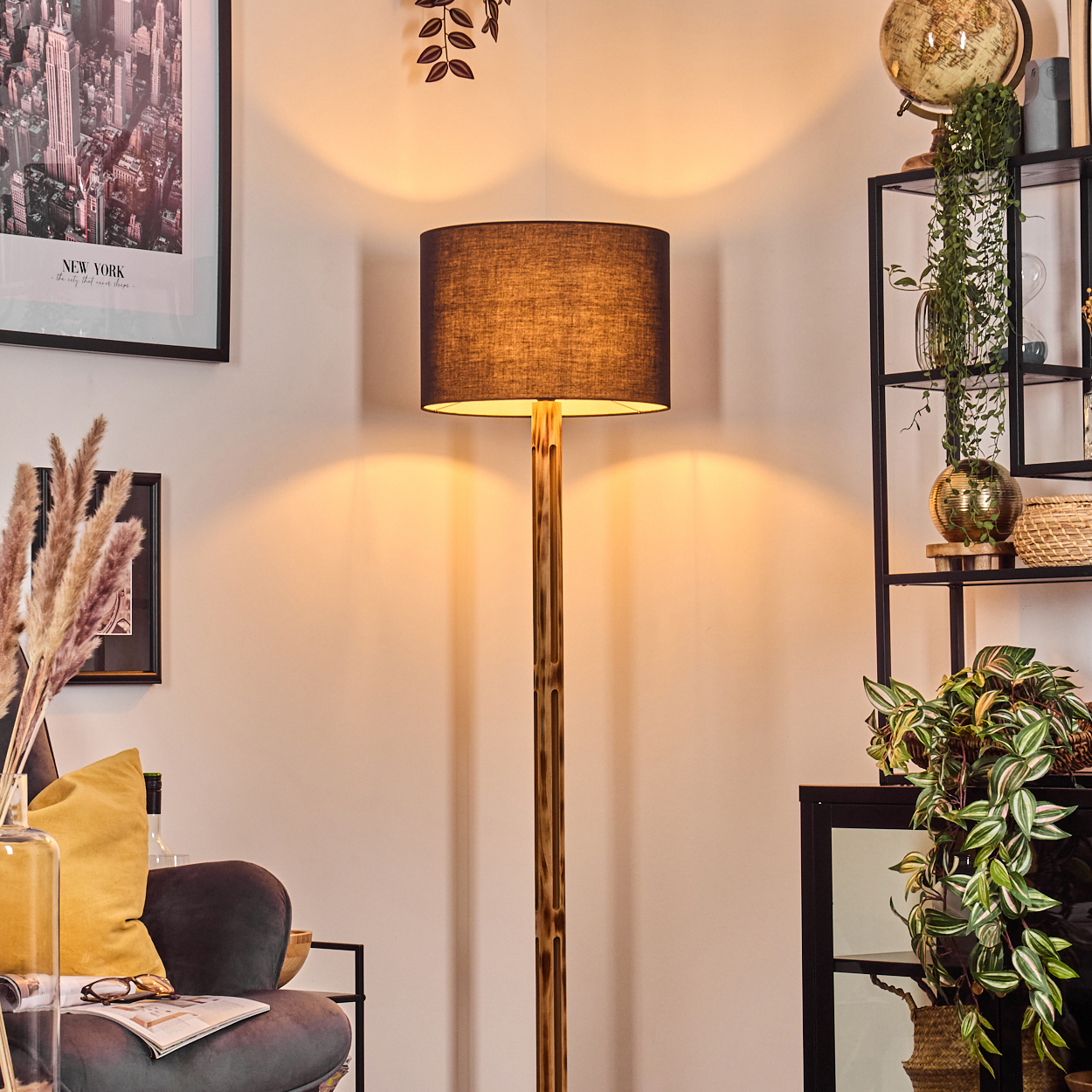 Eine Stehlampe mit schwarzem Lampenschirm, goldener Stange und einem Raum mit Regal, Pflanzen und New York-Poster.
