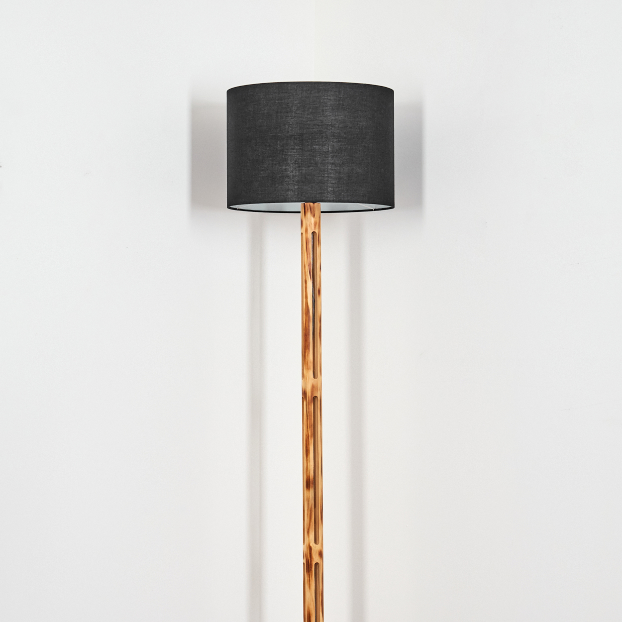 Eine Stehlampe mit schwarzem Lampenschirm und Holzstange steht vor einer weißen Wand. Die Lampe steht in der Ecke des Raumes.