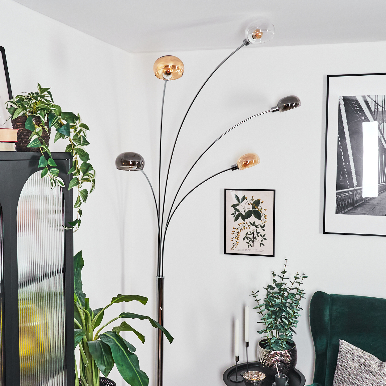Ein modernes Wohnzimmer mit Stehlampe, Pflanzen und gerahmten Bildern. Im Hintergrund ein grüner Sessel.